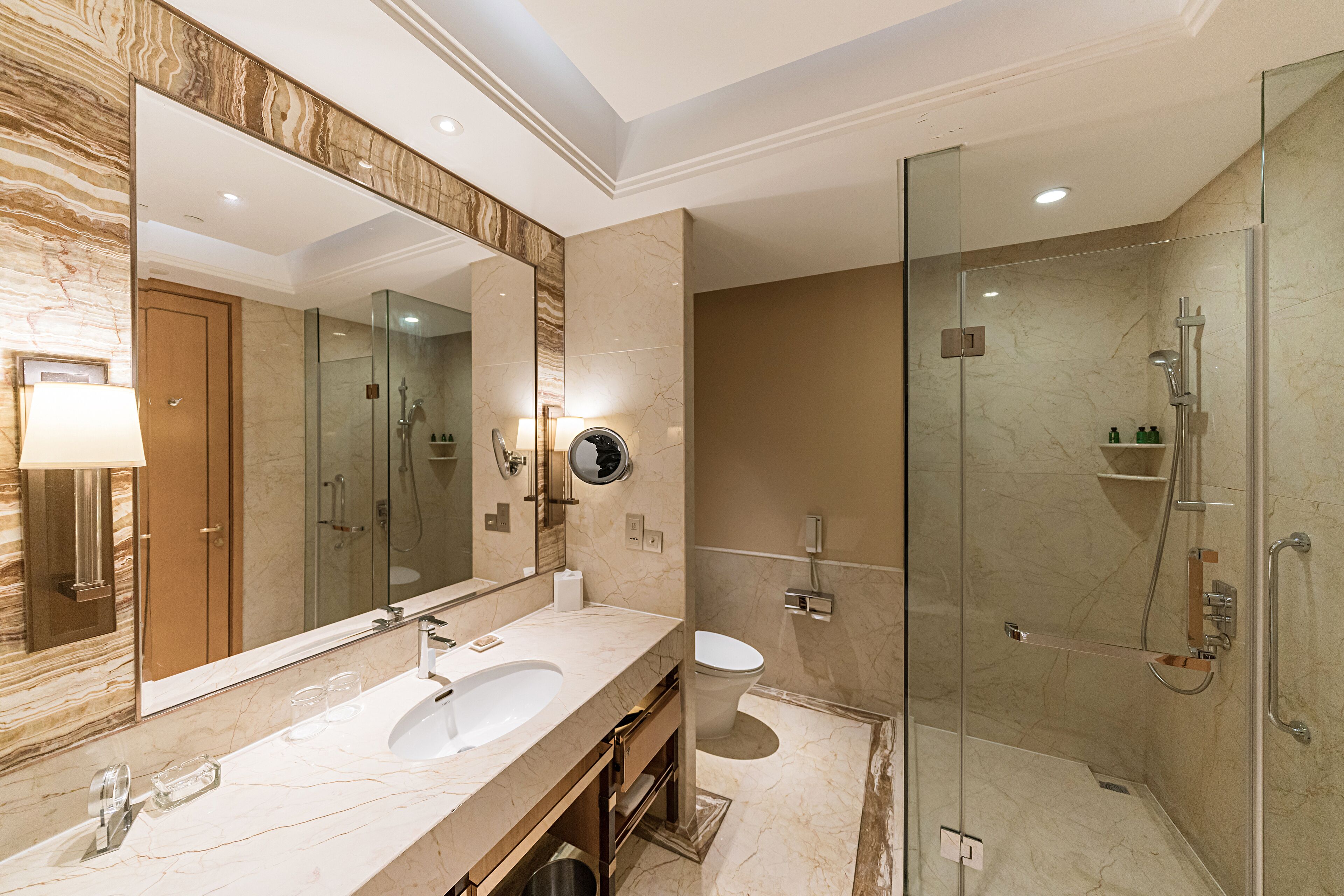A modern bathroom with a large mirror, dual sinks, a glass-enclosed shower, and marble walls.