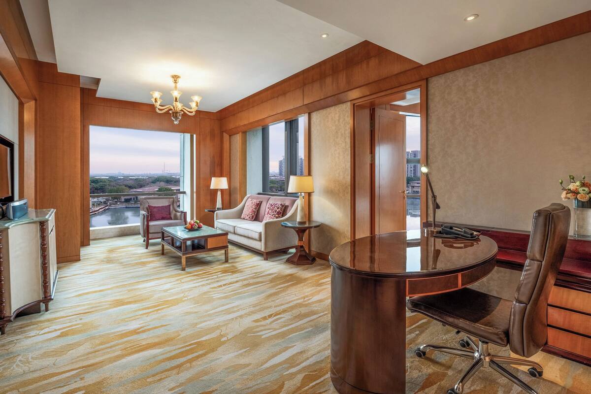 Una habitación de hotel con un ventanal grande, una araña, un escritorio, un sillón de cuero y un sofá.