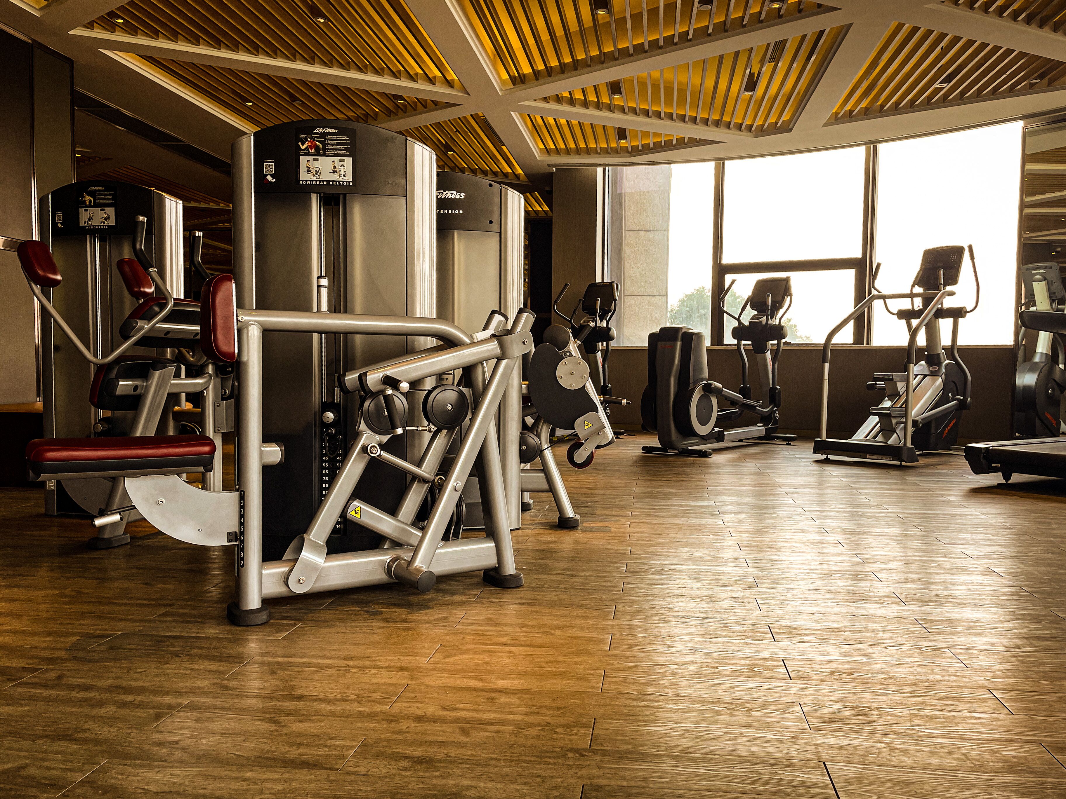 A gym with various exercise machines, large windows, and a wooden floor.