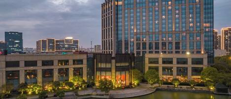 A modern hotel with a large glass facade, a courtyard with trees, and a body of water in front.
