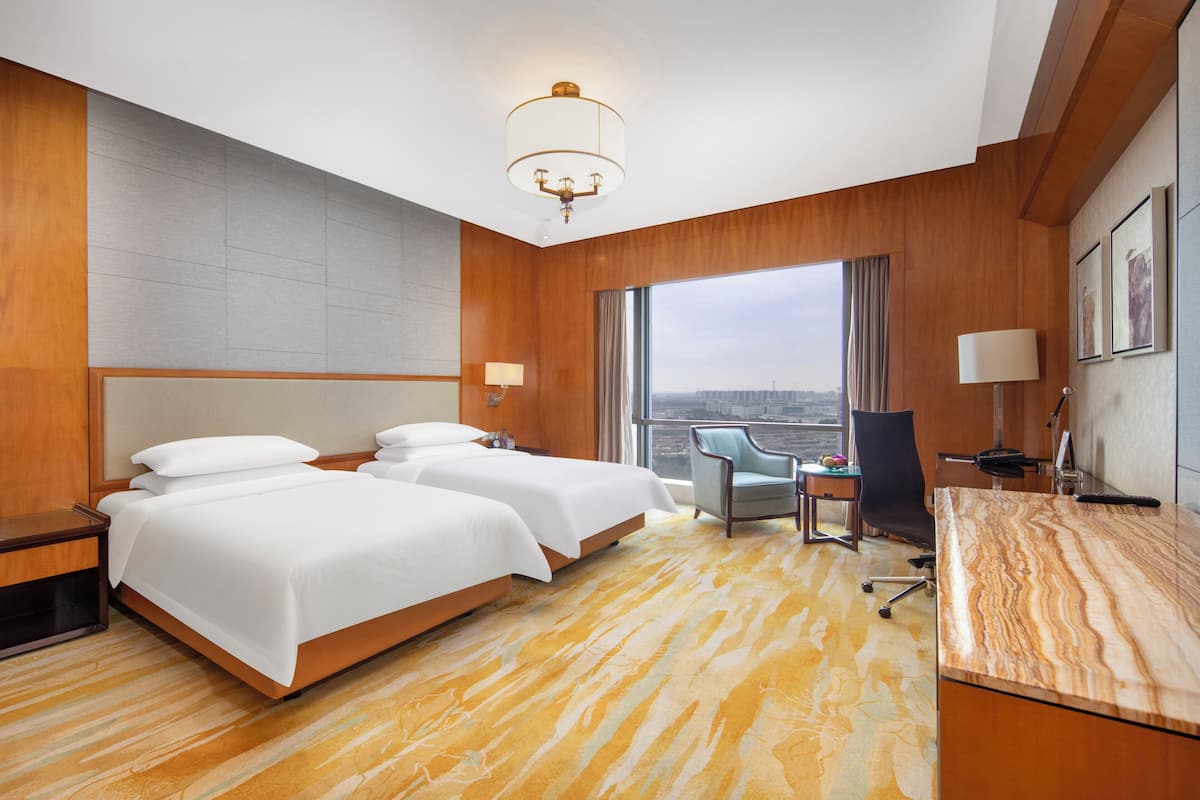 Habitación de hotel con dos camas, un escritorio, una silla, una lámpara y un amplio ventanal con vista a la ciudad.