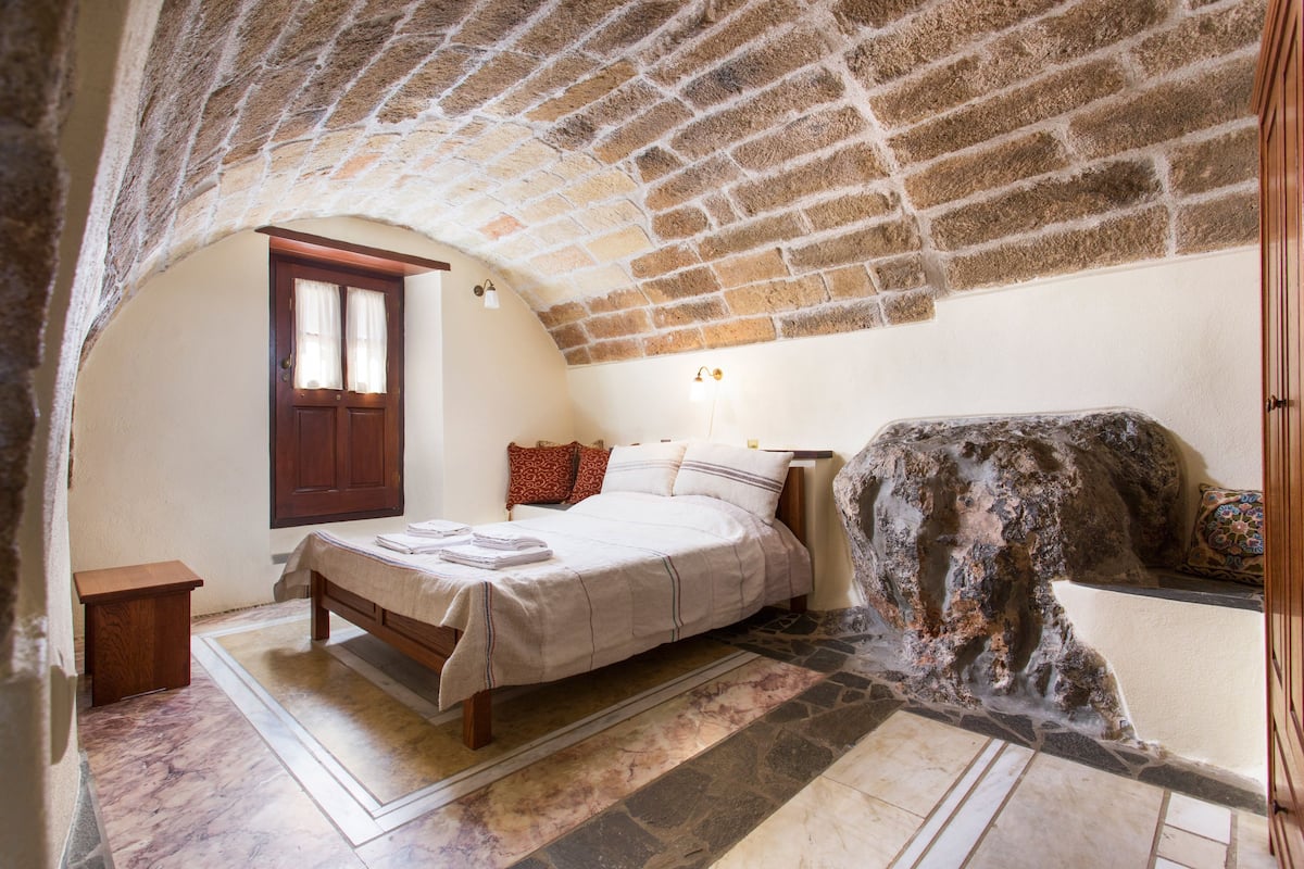 A bedroom with a stone ceiling, a bed with white linens, a small wooden nightstand, and a stone wall.
