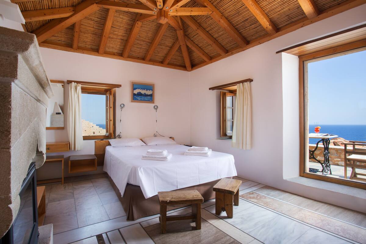 A bedroom with a large bed, wooden benches, a desk, and a view of the sea.