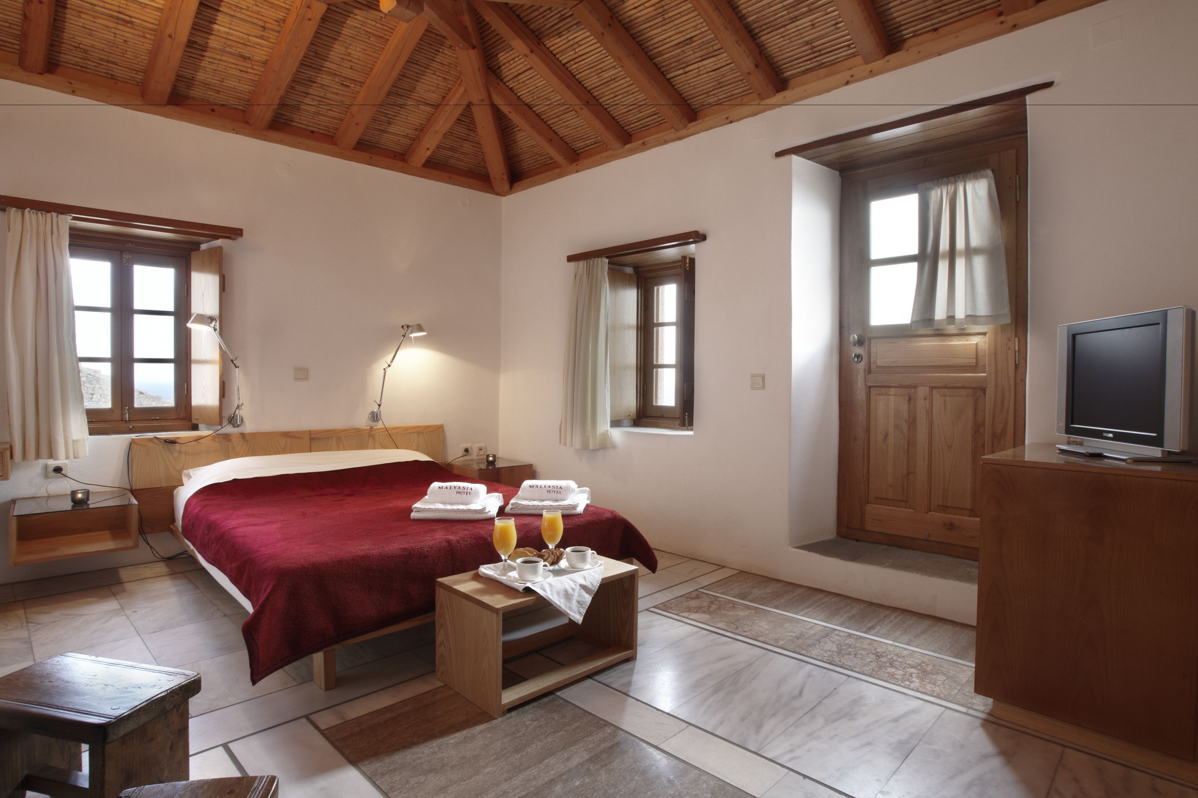 A hotel room with a wooden ceiling, a bed with a red blanket, a bedside table with glasses and a towel, a television on a wooden stand, and a window with curtains.