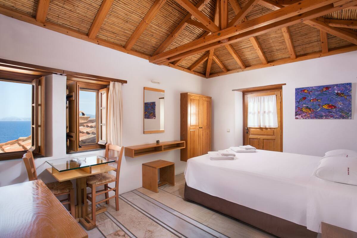 A bedroom with a wooden ceiling, a bed, a desk with a chair, a wardrobe, and a painting on the wall.