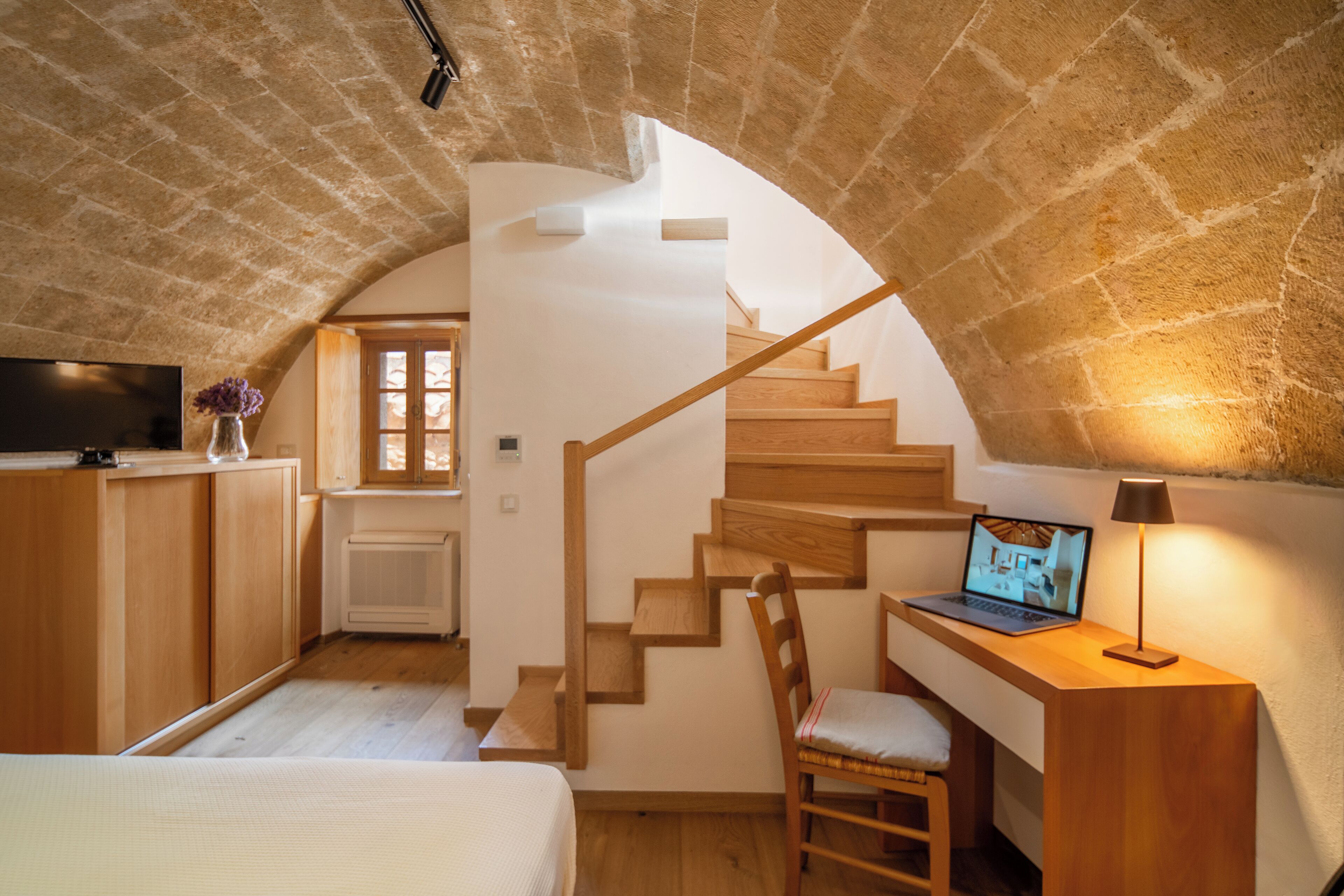 A cozy room with a wooden desk, a laptop, and a chair. There's a staircase with wooden steps leading to an upper level.