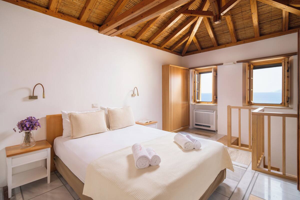 A bedroom with a large bed, wooden ceiling beams, a small table with a vase of flowers, and a view of the sea through a window.