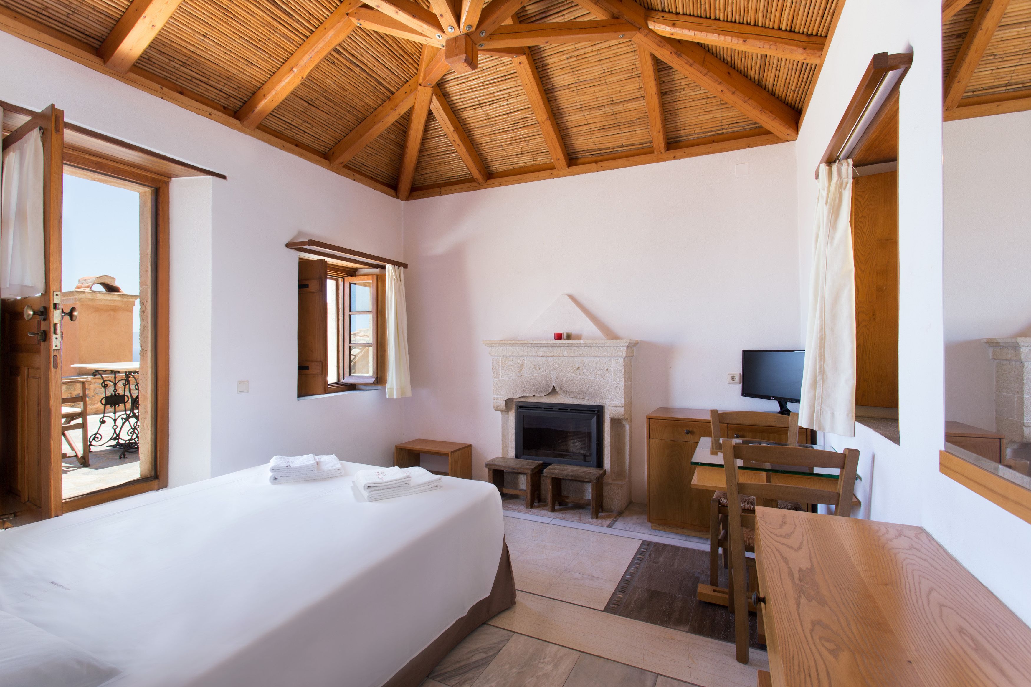 A bedroom with a large bed, wooden ceiling beams, a fireplace, a television, and a balcony visible through an open door.
