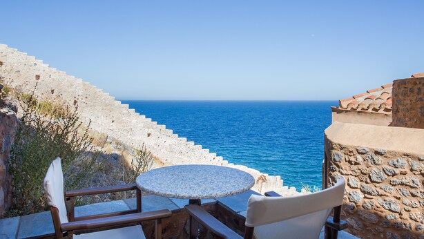 Un escalier en pierre mène à une terrasse où se trouvent une table ronde et deux chaises, d’où l’on peut admirer une mer d’un bleu limpide.