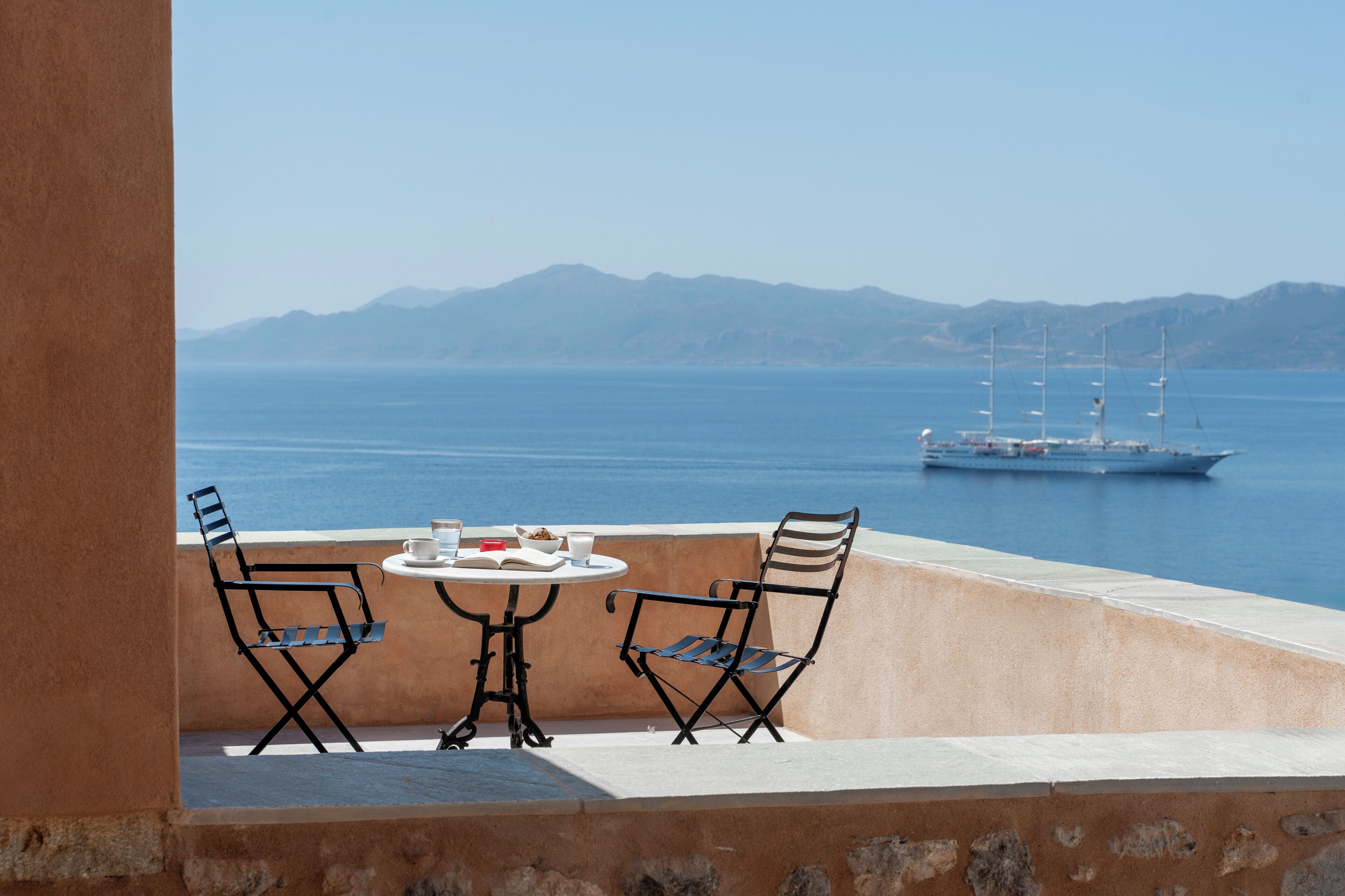 A terrace with two chairs and a table, overlooking a calm sea and distant mountains.