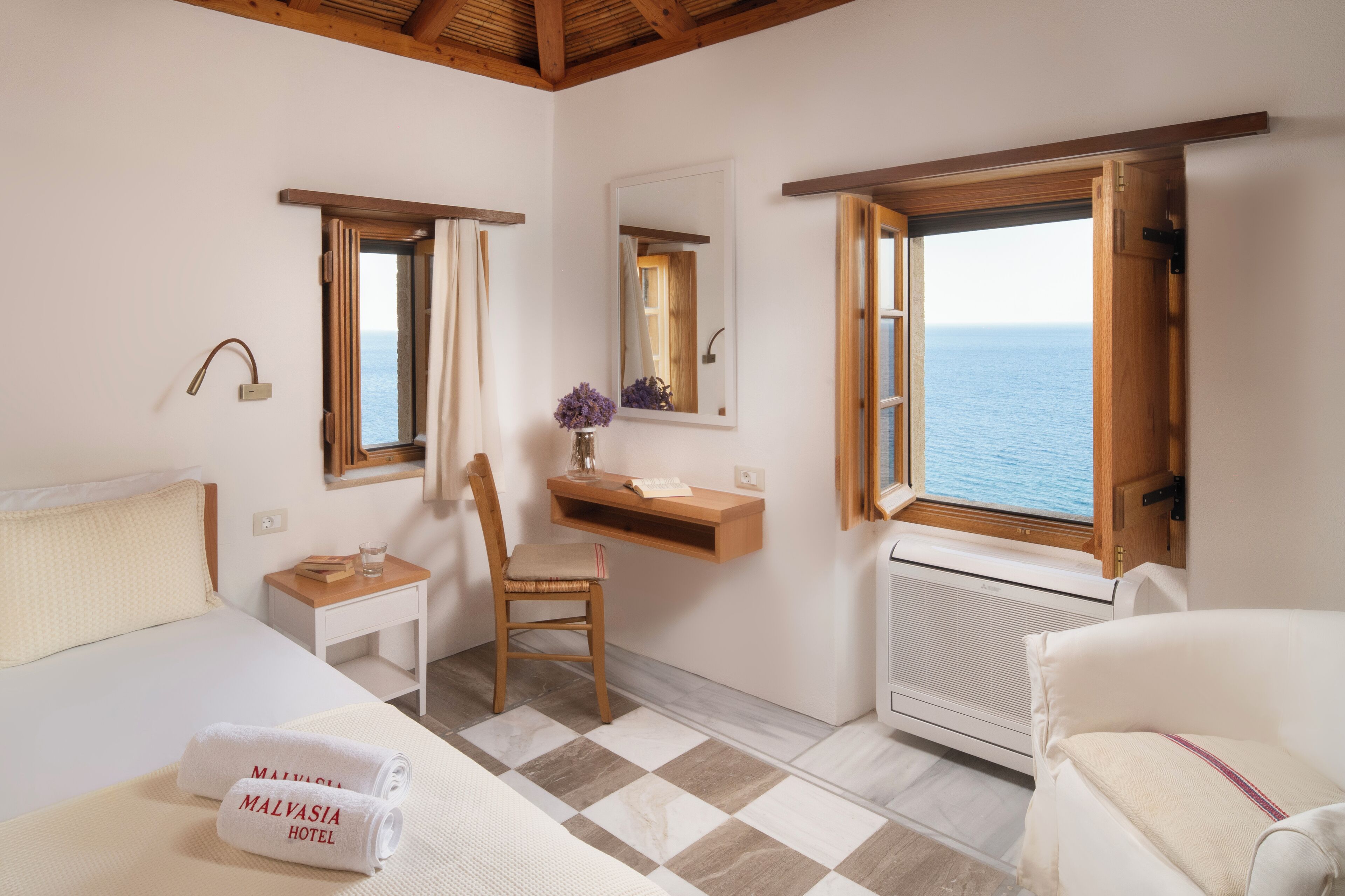A hotel room with a bed, a chair, a small table, a mirror, and a window with a view of the sea.