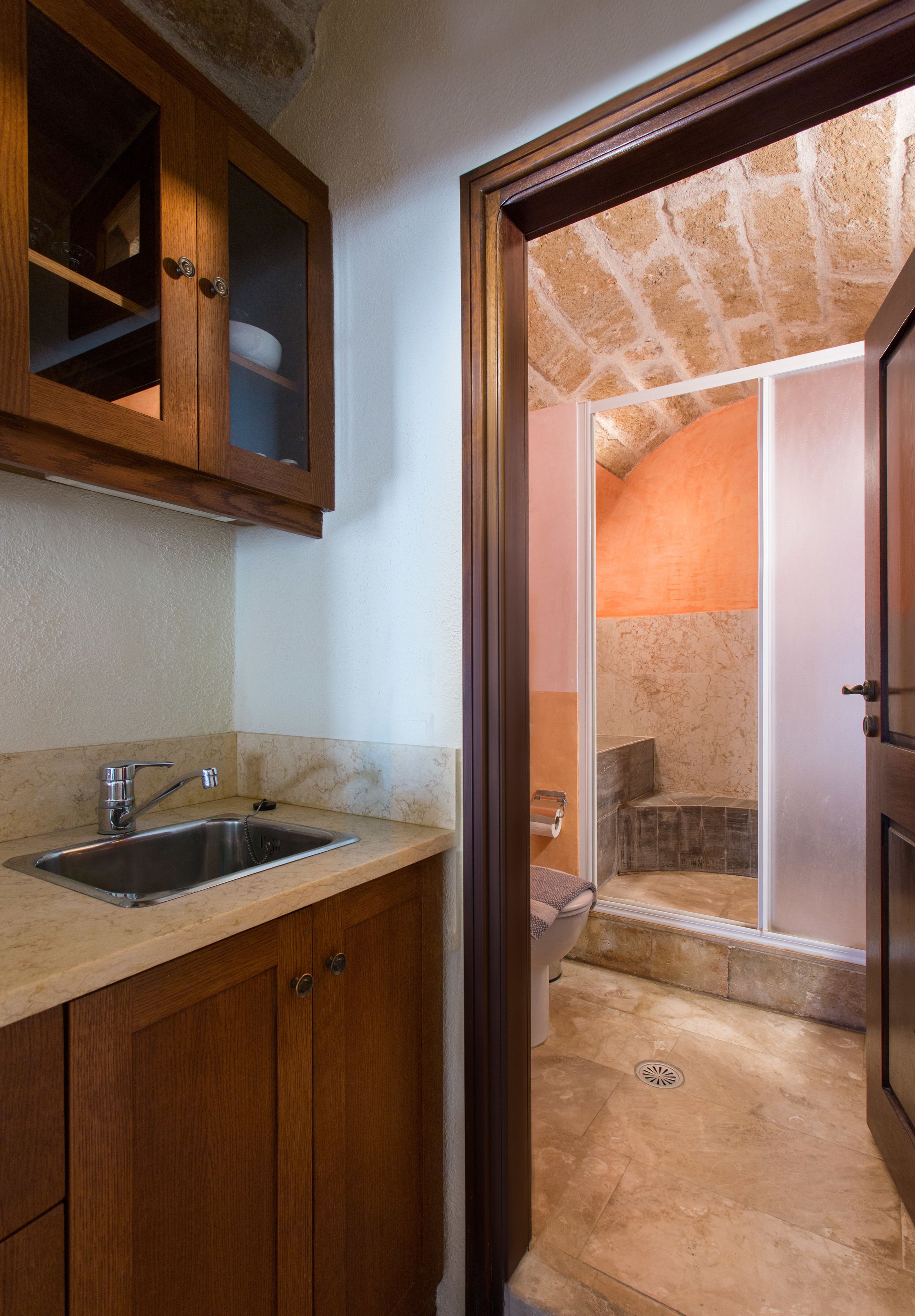 Cocina con armarios de madera y un lavabo, que da acceso a un baño con ducha y suelo de mármol.
