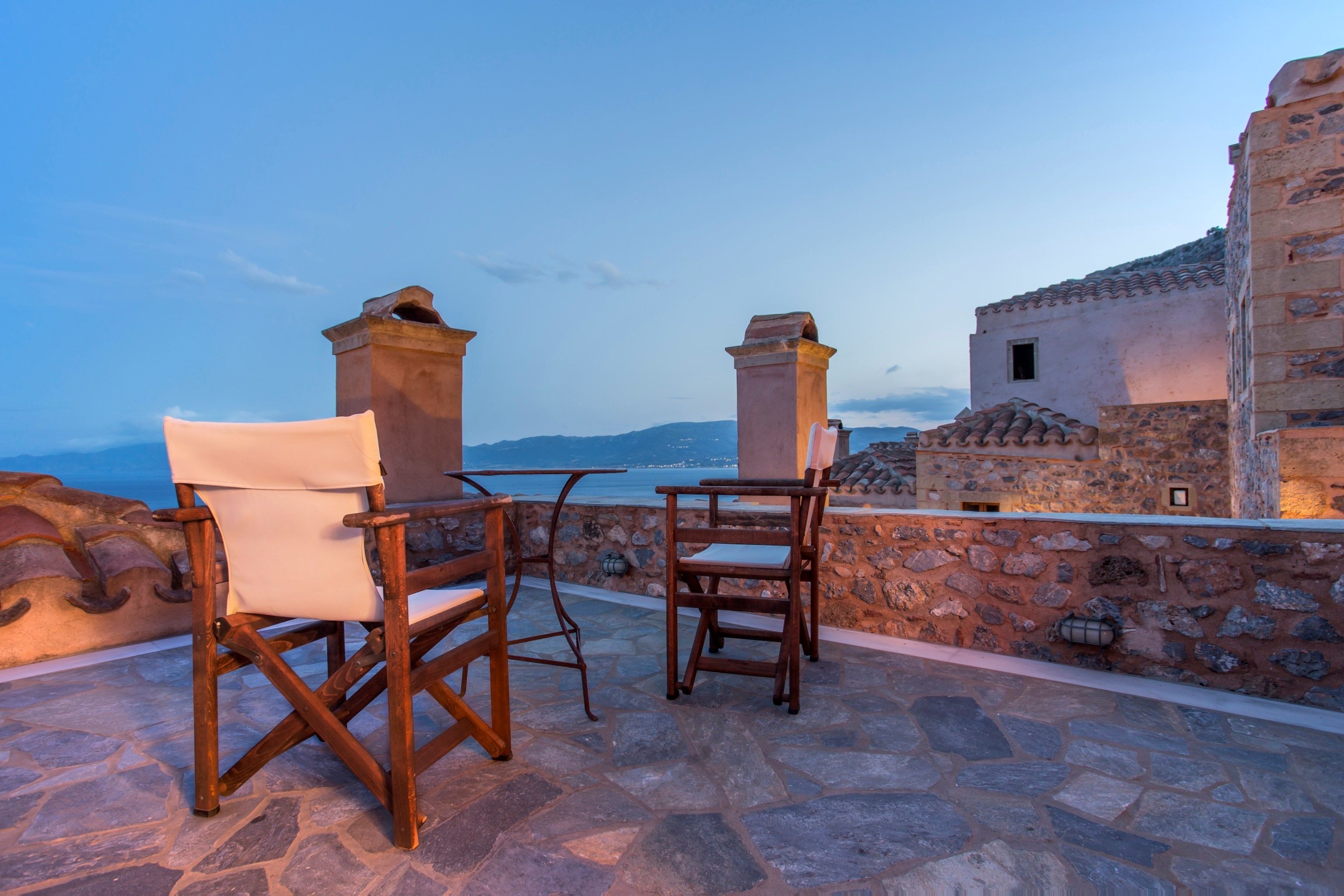 A stone patio with two wooden chairs and a table overlooking a coastal view.