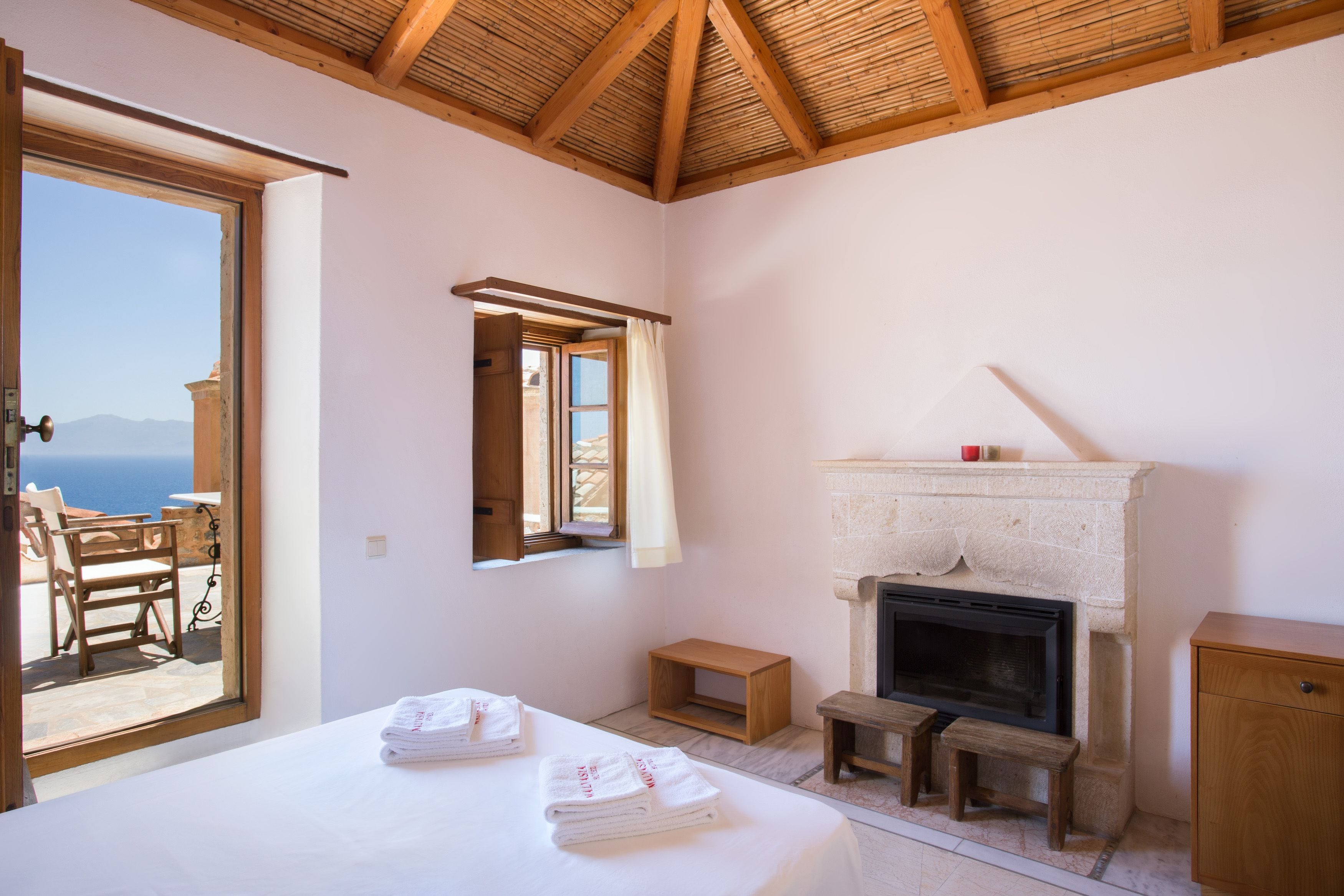 A bedroom with a bed, a fireplace, a wooden bench, and a view of the sea through an open door.