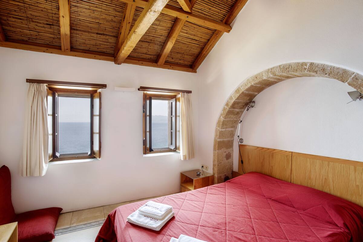 A bedroom with a wooden ceiling, two windows with curtains, a red bed, and a stone archway.