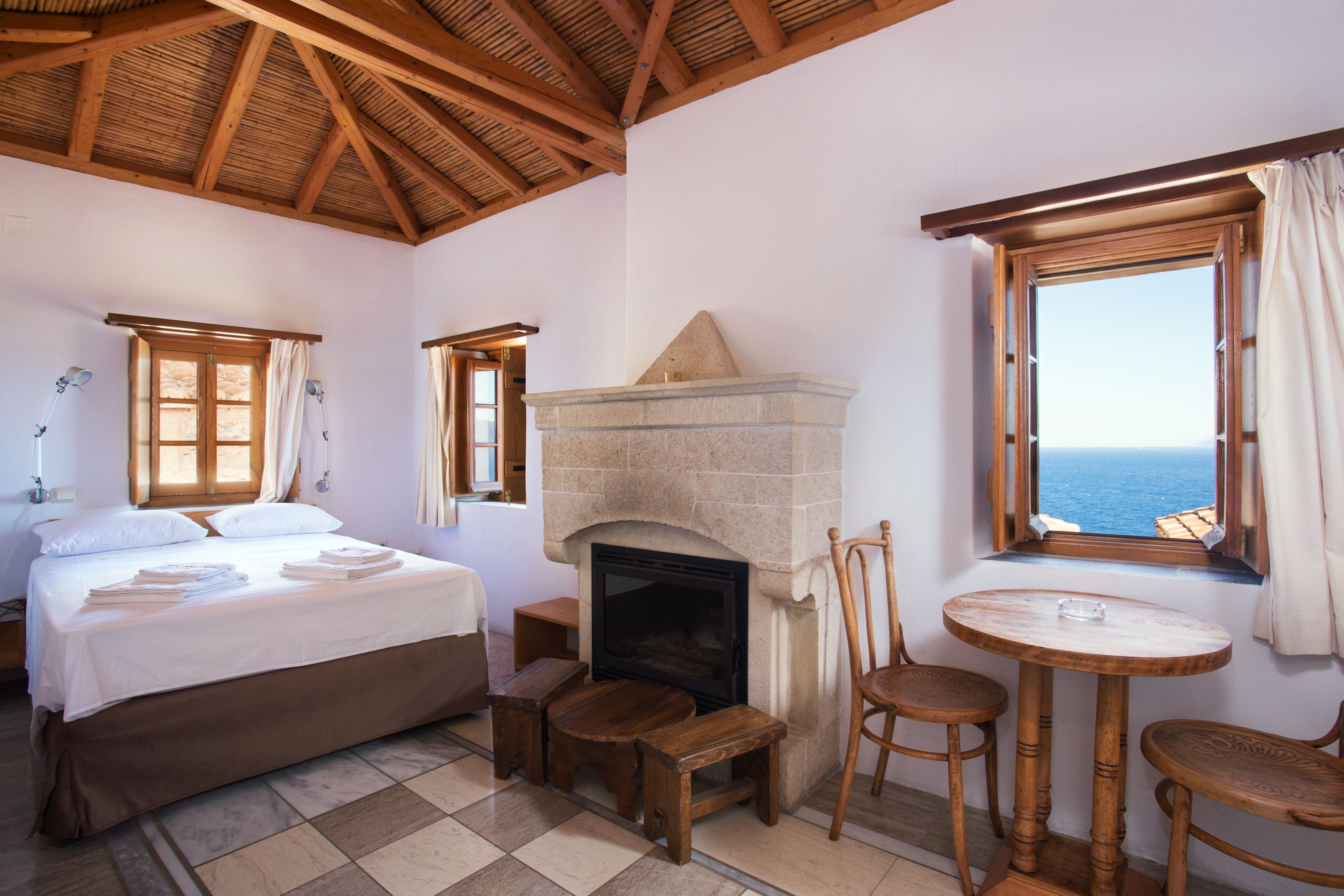 A bedroom with a bed, a fireplace, a table with chairs, and a view of the sea.