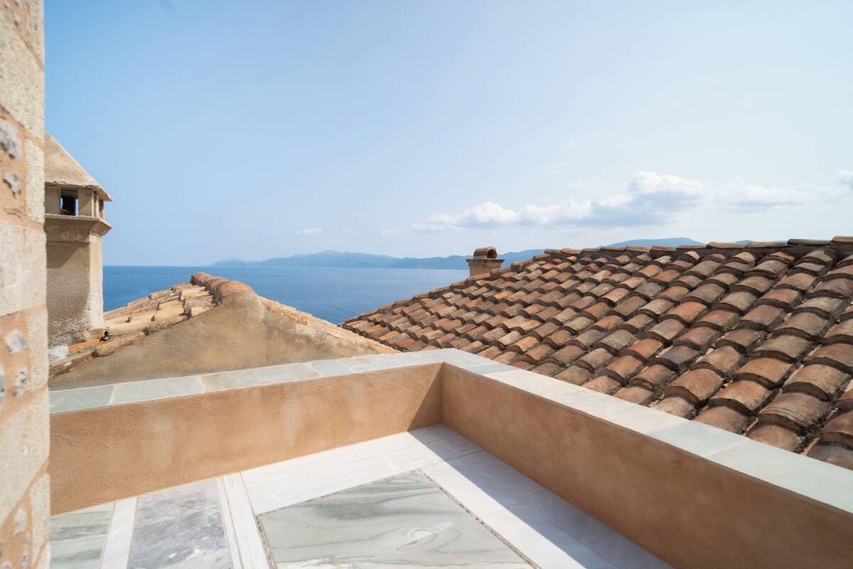 A terrace with a view of the sea and distant mountains, featuring terracotta roof tiles and a stone wall.