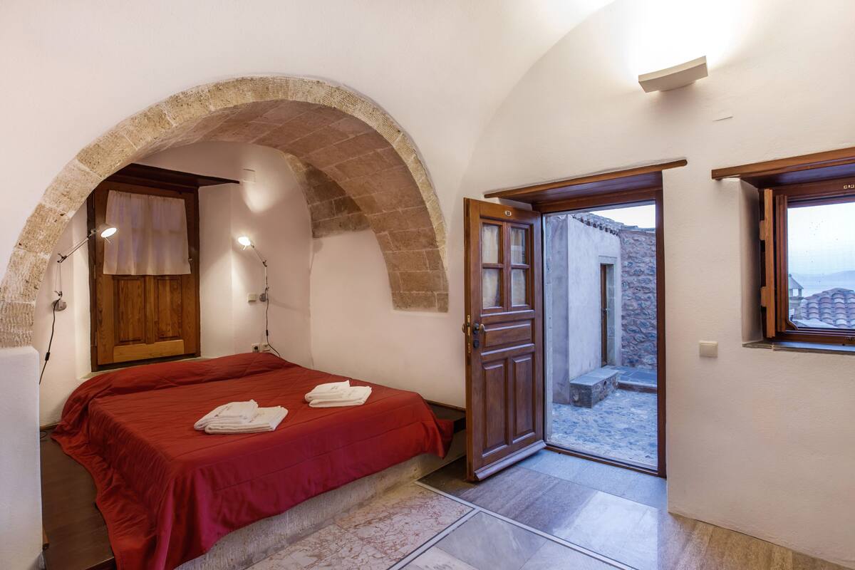 A bedroom with a red bedspread, an arched doorway, a wooden door, and a view of the outside.