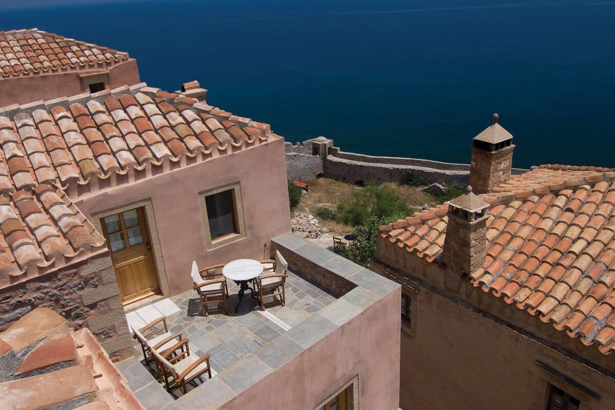 A building with terracotta-tiled roofs, a stone wall, and a small table with chairs on a flat roof area.