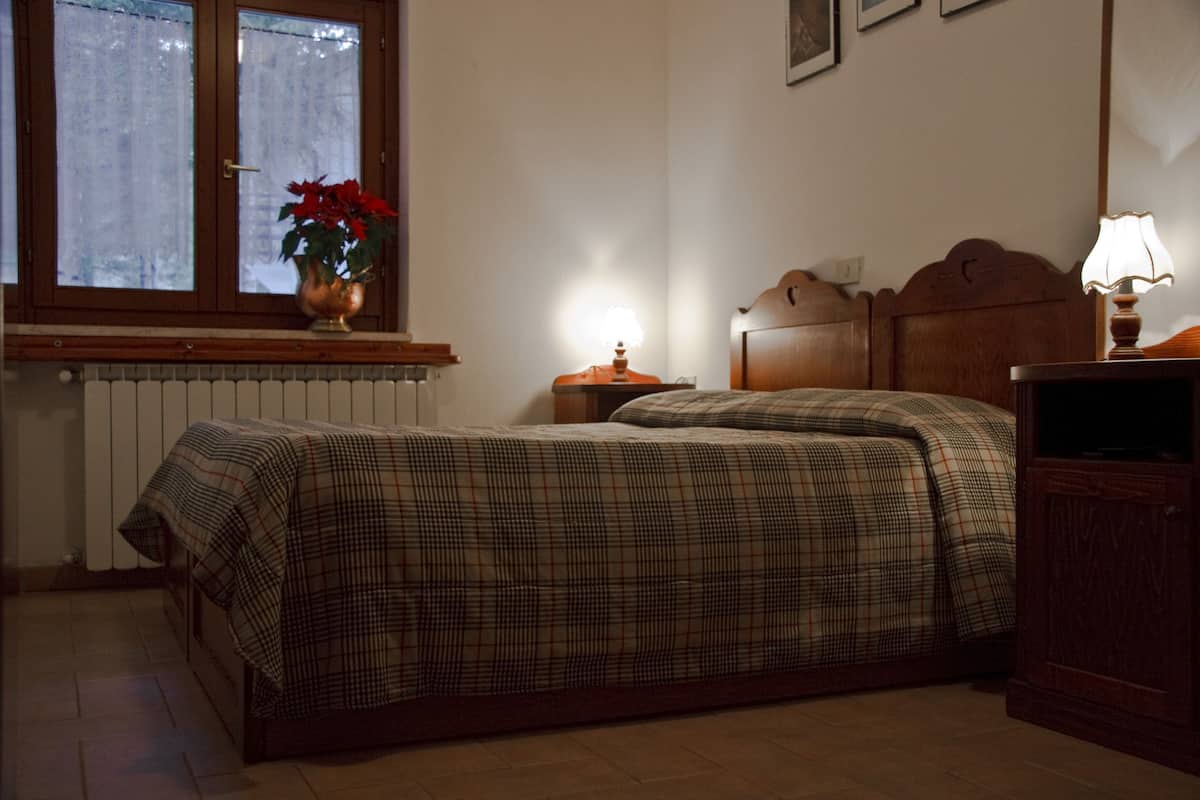 A bedroom with a bed, a nightstand, a lamp, a window with curtains, and a radiator.