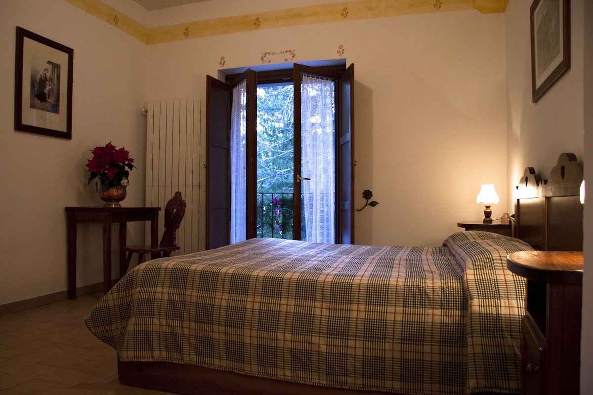 Quarto com cama, janela com cortinas, mesa de cabeceira, abajur e um quadro na parede.
