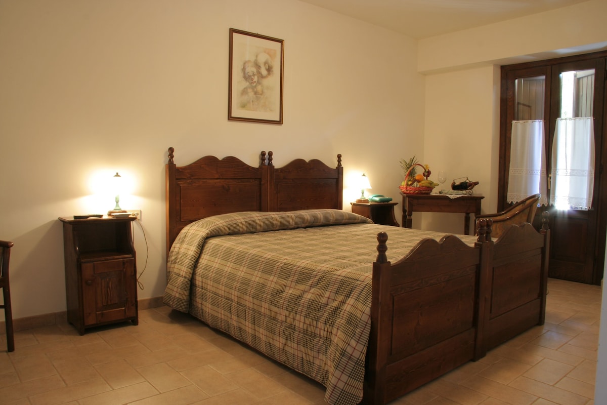 Quarto com cama de madeira, mesas de cabeceira, criadeira e um quadro na parede, além de uma janela com cortinas.