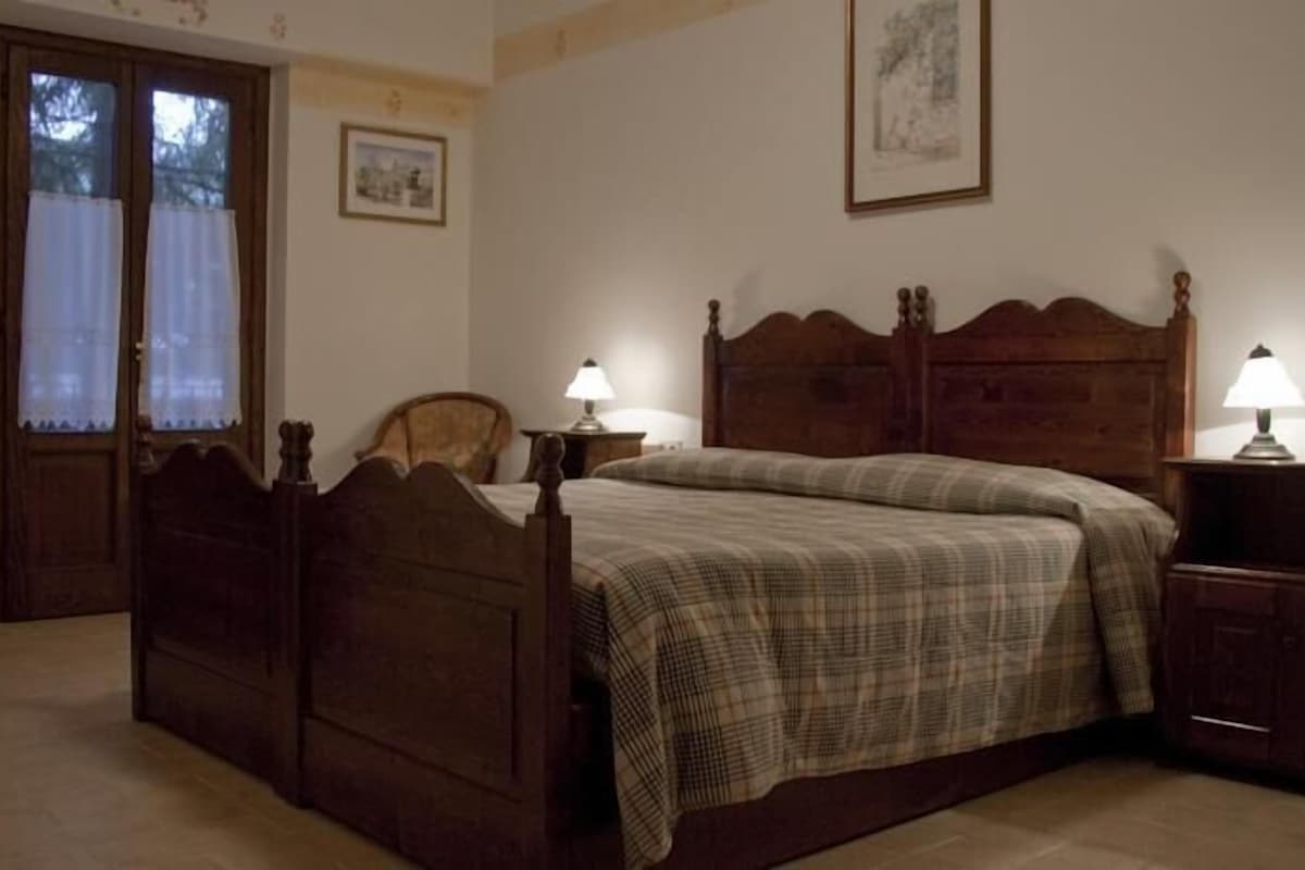 Quarto com cama de madeira, duas mesas de cabeceira com luminárias, uma cadeira, uma porta e um quadro na parede.