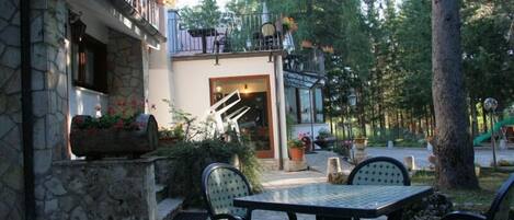 An outdoor patio with stone walls, a glass table, and chairs. There is a staircase leading to a balcony with outdoor seating.