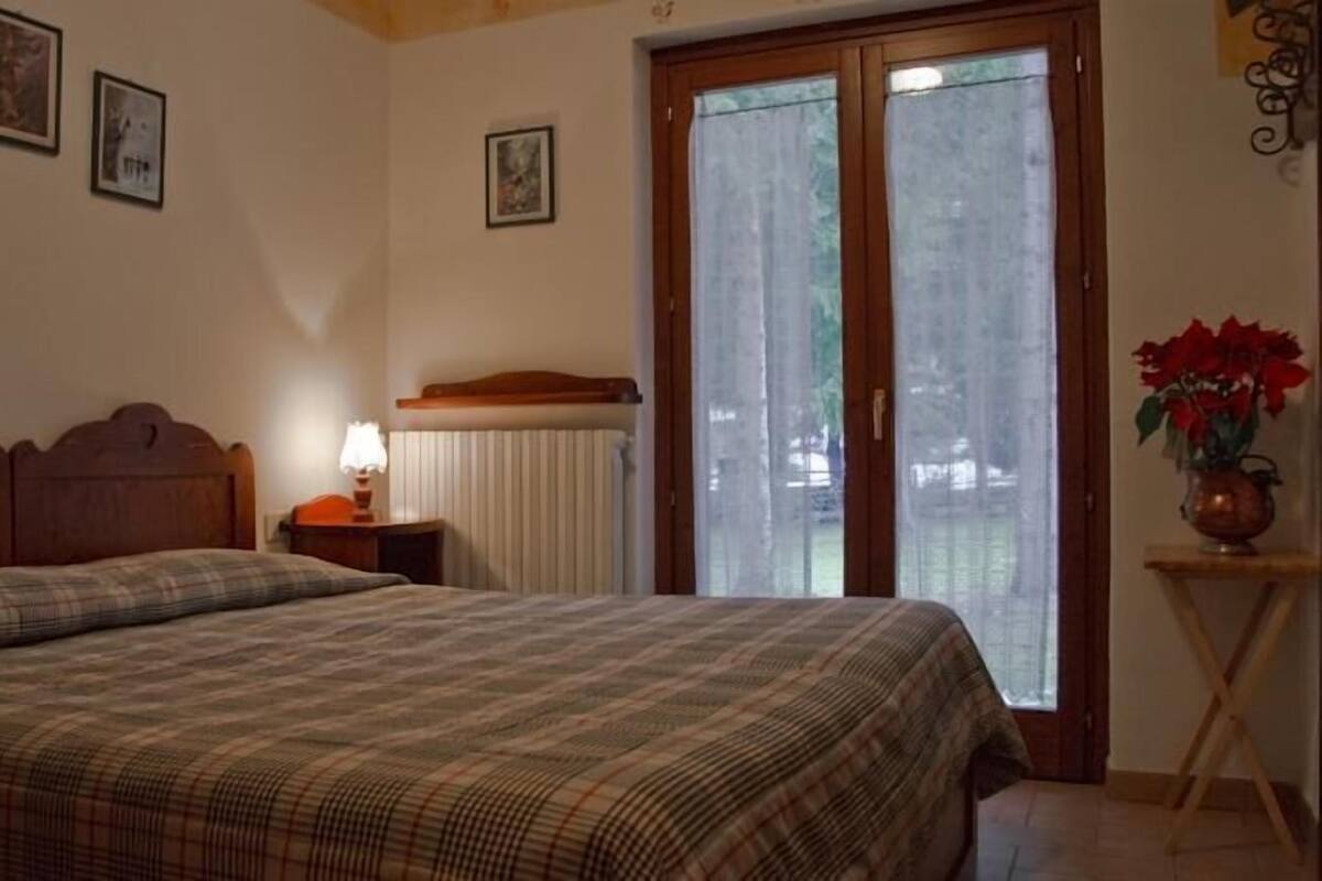 A bedroom with a double bed, a bedside table with a lamp, a radiator, and a window with curtains.