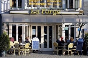 An outdoor seating area of La Forge with wicker chairs and tables, a glass door entrance, and a signboard. - Hotel La Forge (Malmedy)