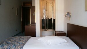 Una habitación de hotel con cama, una mesita de noche pequeña, un armario y zona de ducha.