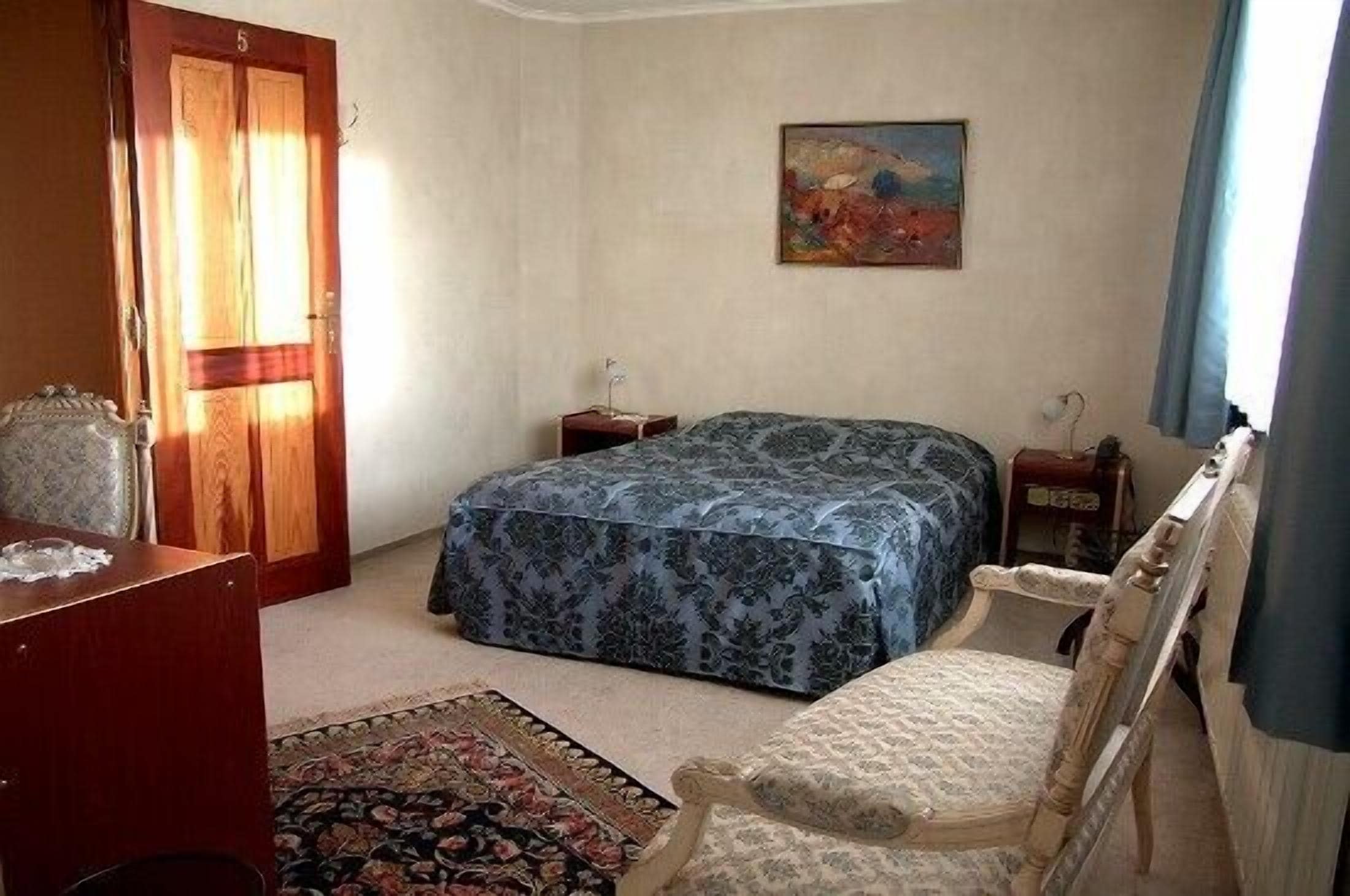 A bedroom with a bed, a chair, a desk, a nightstand, a painting, and a door.