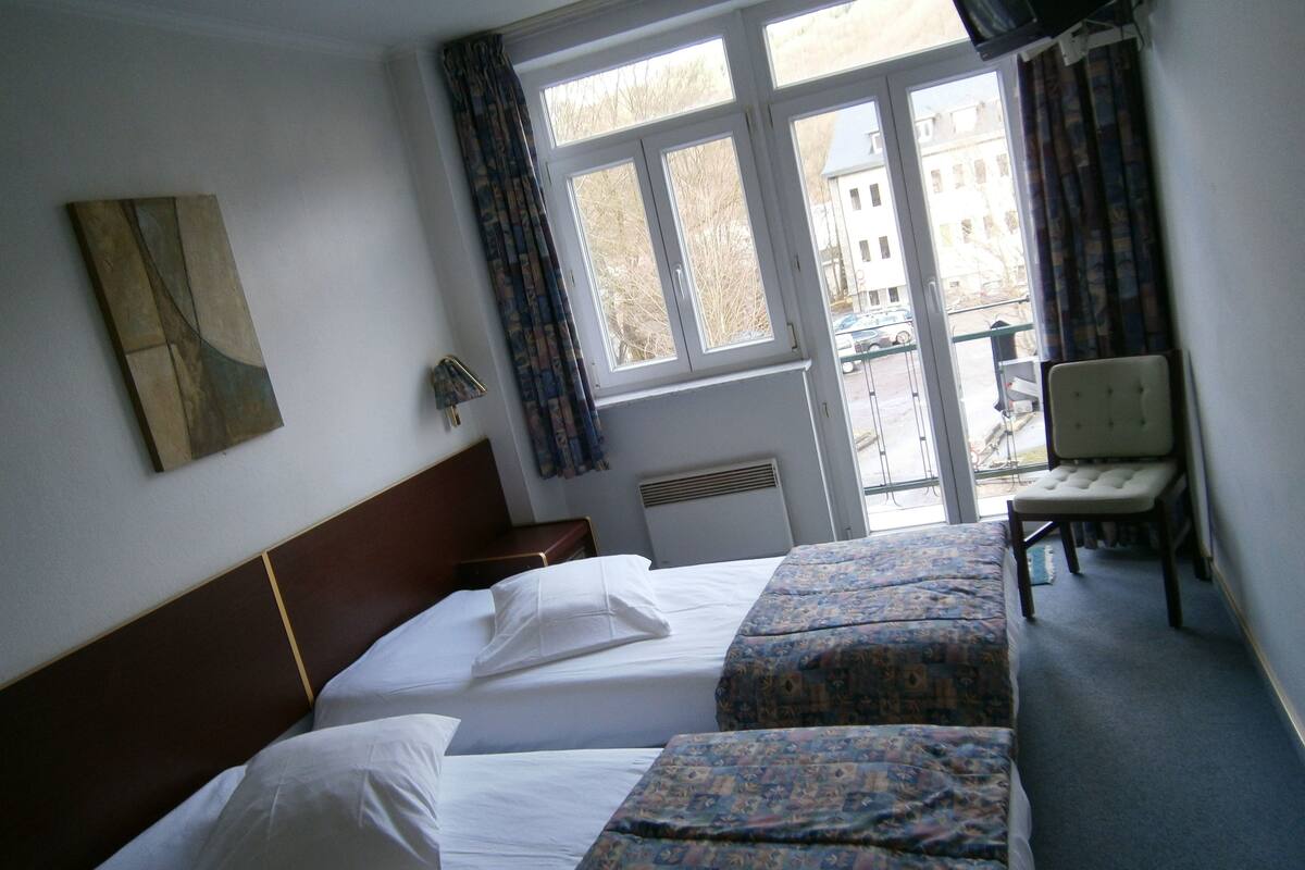 A hotel room with two beds, a chair, a window with curtains, and a radiator.