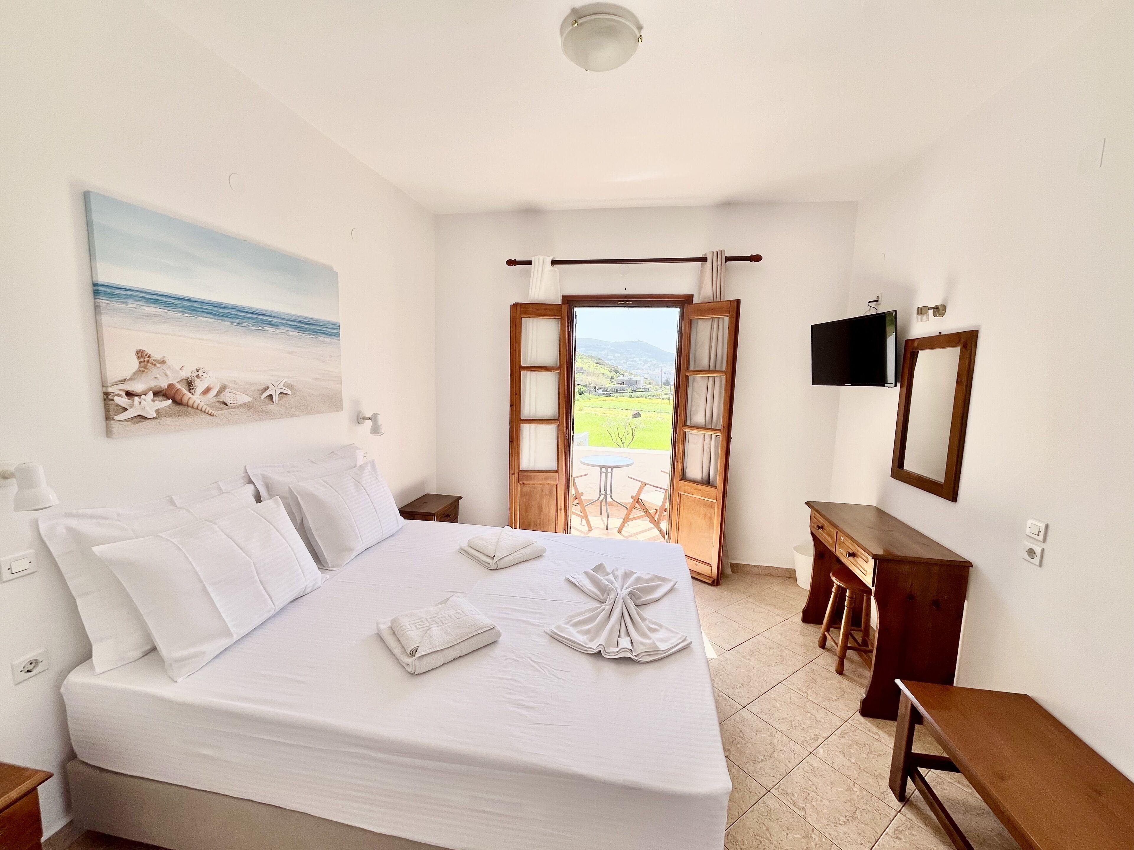 A bedroom with a bed, a desk, a chair, a TV, and a door leading to a balcony with a view of the countryside.