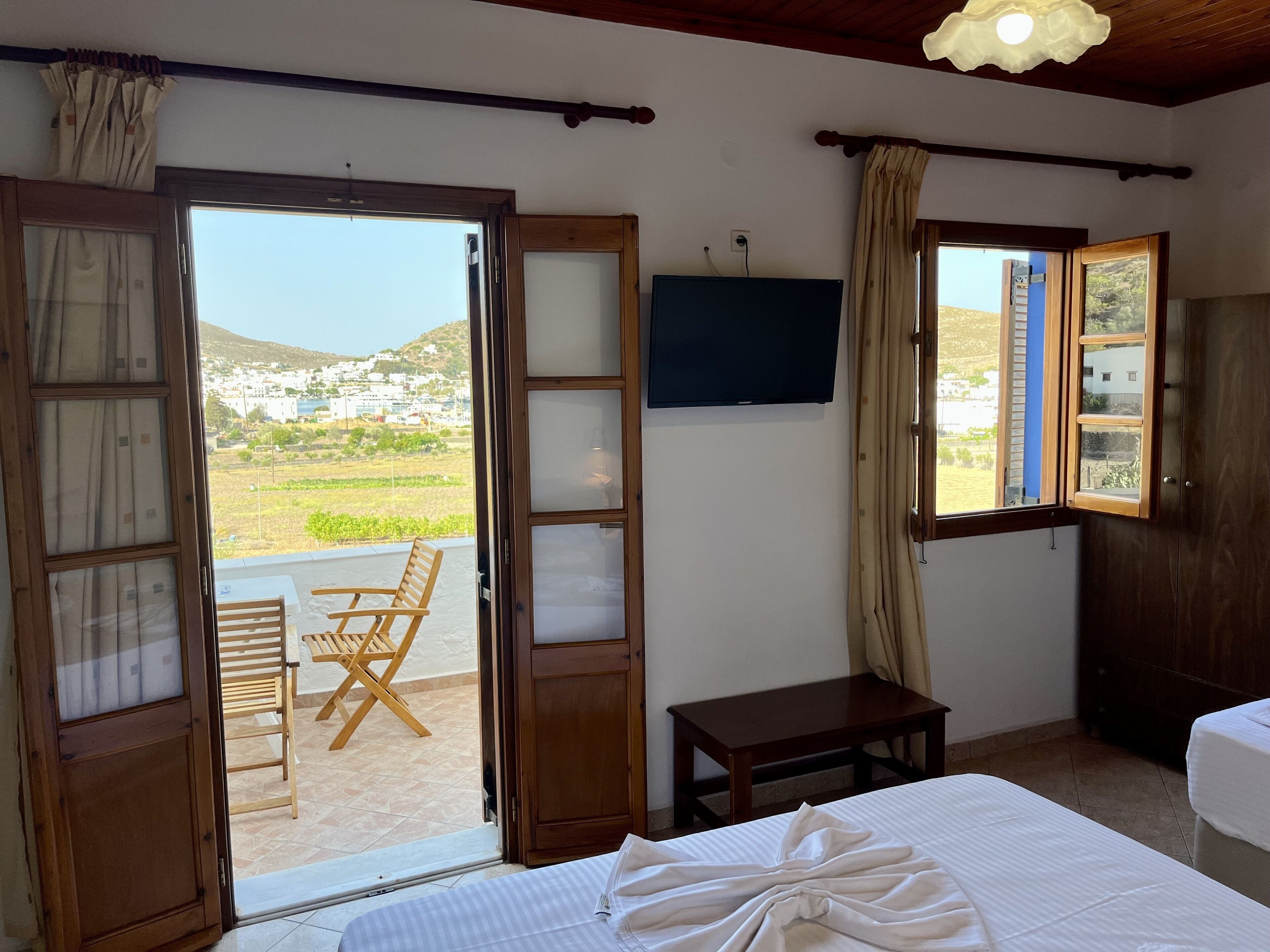 A room with a bed, a TV, a small table, and a chair. There is a window with curtains and a view of a landscape.