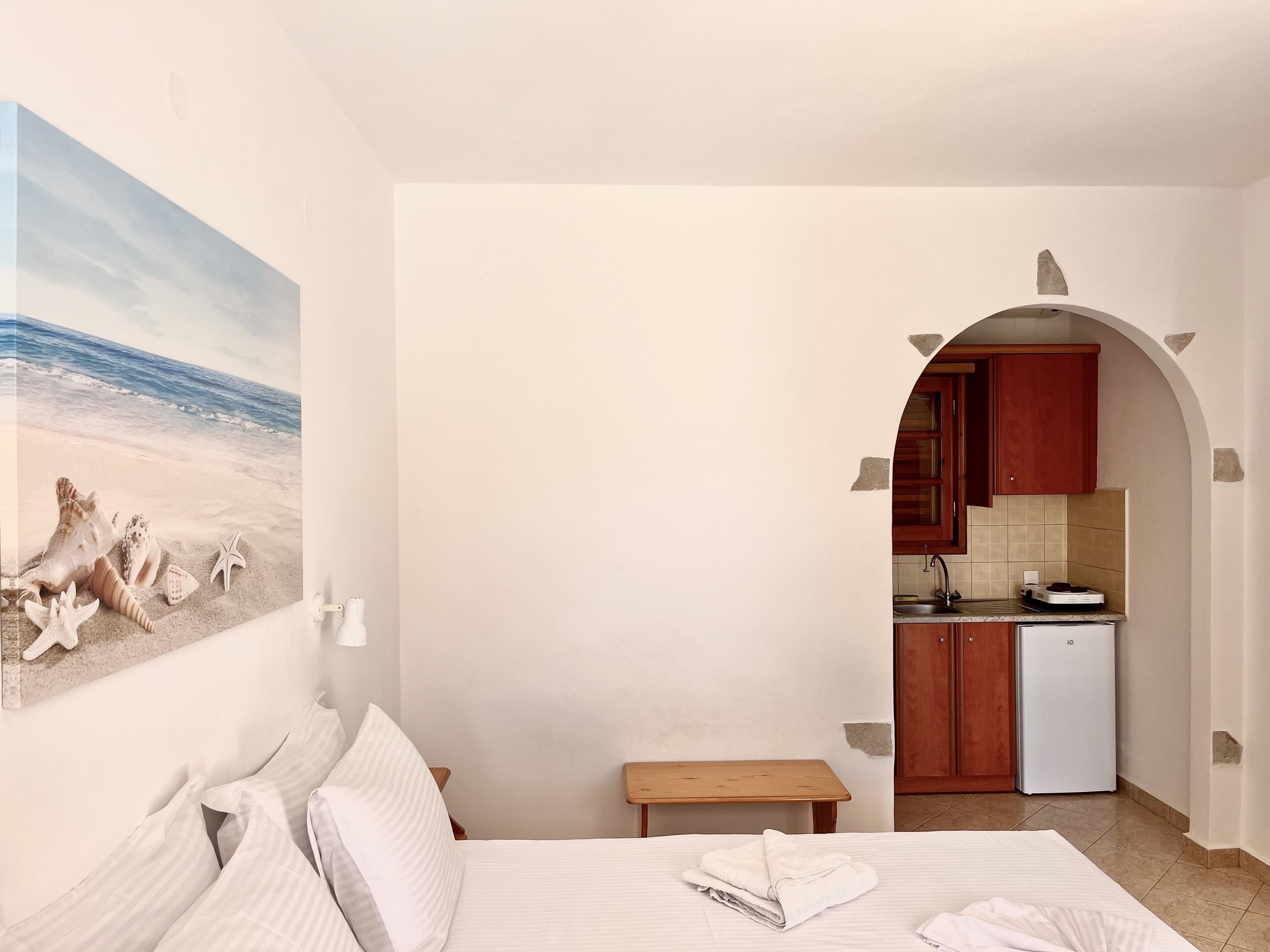 Una camera da letto con un letto, cuscini, un quadro raffigurante una spiaggia, una piccola panca di legno e vista su una cucina con lavandino e frigorifero.