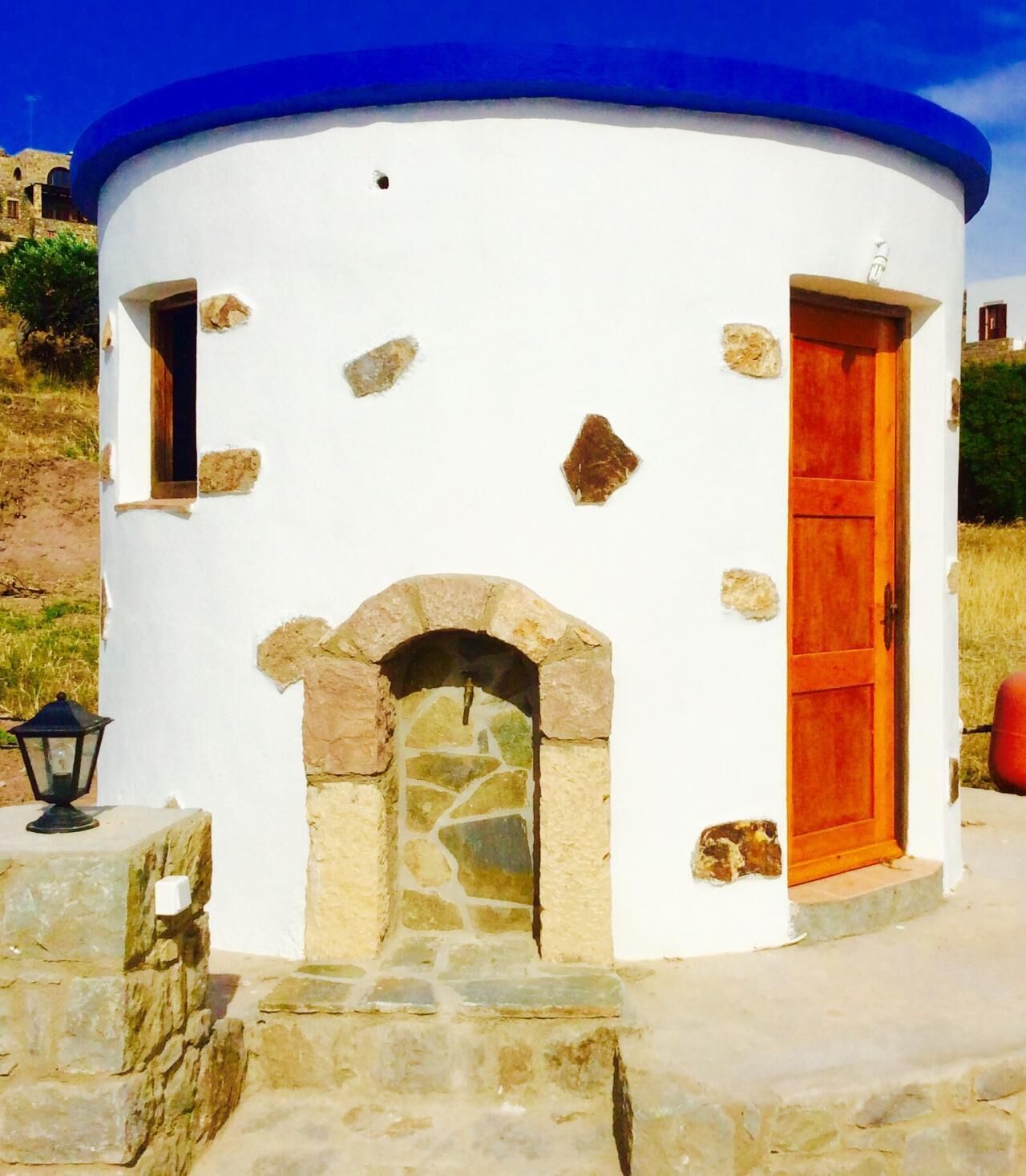 Un petit bâtiment blanc, avec un toit bleu, une entrée en forme d’arc en pierre et une porte rouge.