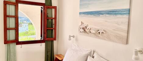 Un dormitorio con cama, almohadas, una mesita de noche, una ventana con cortinas y un cuadro con motivos de playa en la pared.