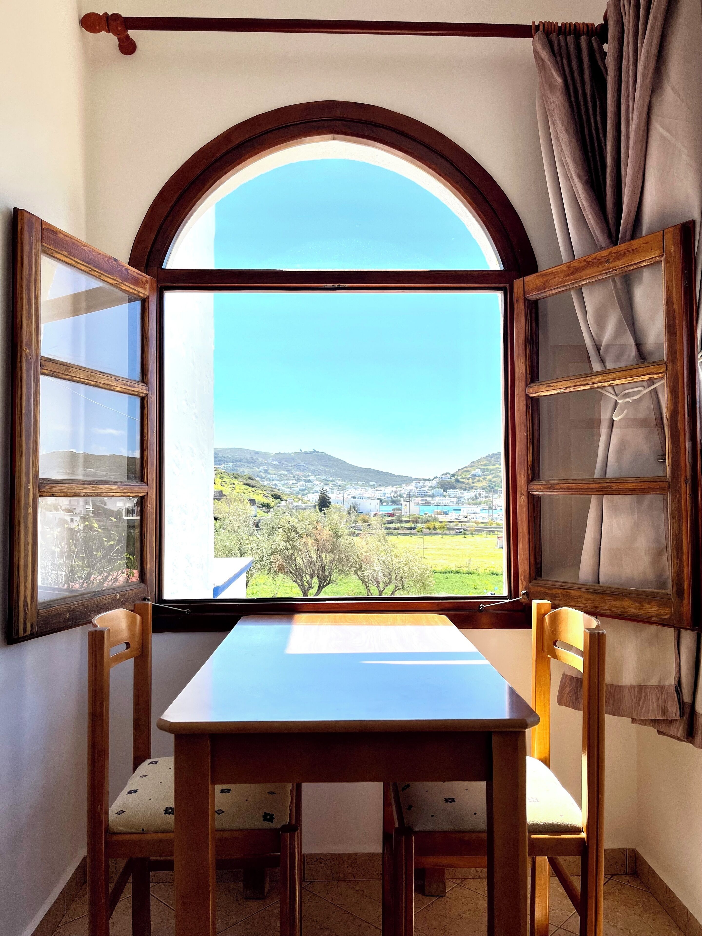 A room with a table and two chairs, a window with curtains, and a view of a landscape with buildings and trees.
