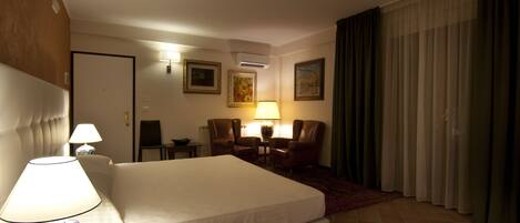 A hotel room with a large bed, two chairs, a table, a lamp, and a painting on the wall.