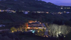 A large house with a swimming pool surrounded by trees, set on a hillside with other houses in the background.