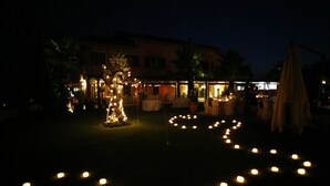A nighttime outdoor area with a pathway of lit candles, a small tree decorated with lights, and a building in the background.