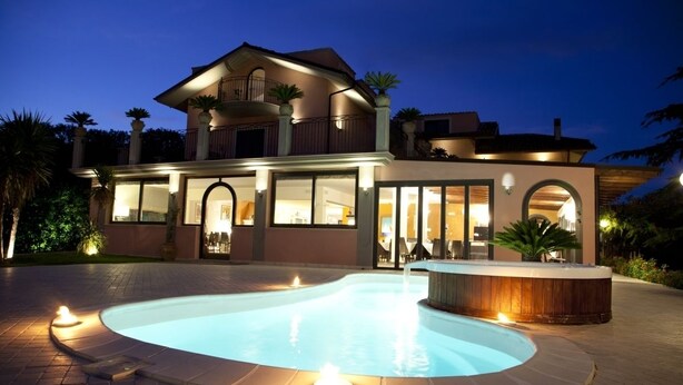 A large, two-story house with a swimming pool and hot tub, surrounded by well-lit outdoor areas and palm trees.