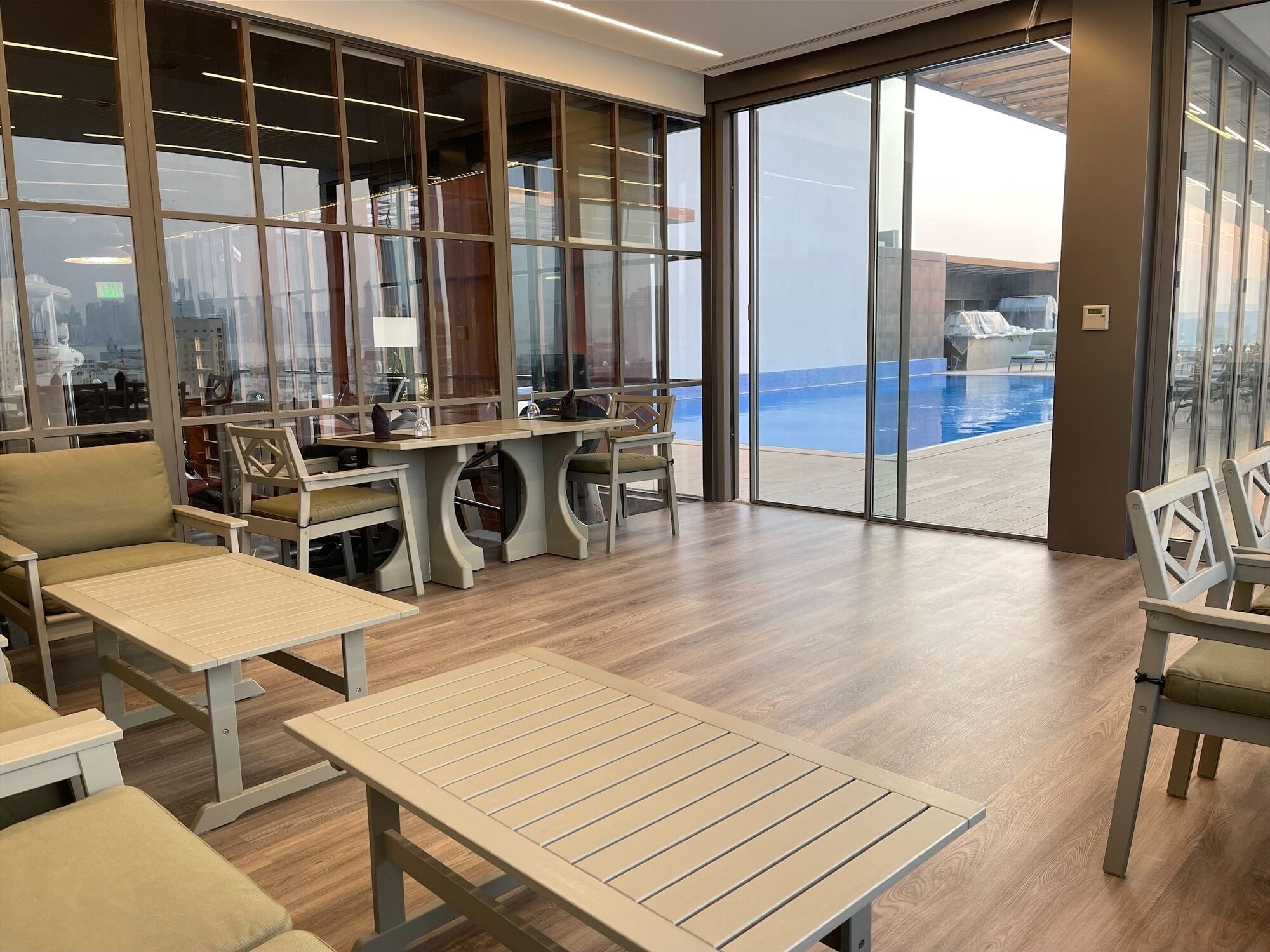 Espaço moderno interno com piso de madeira, mesas e cadeiras. Grandes portas de vidro dão acesso a uma área de piscina externa.