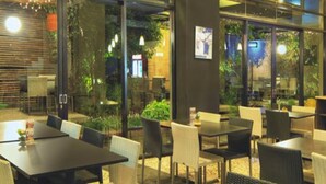 Un área de comedor al aire libre con mesas y sillas, ventanas amplias y follaje visible a través del vidrio.