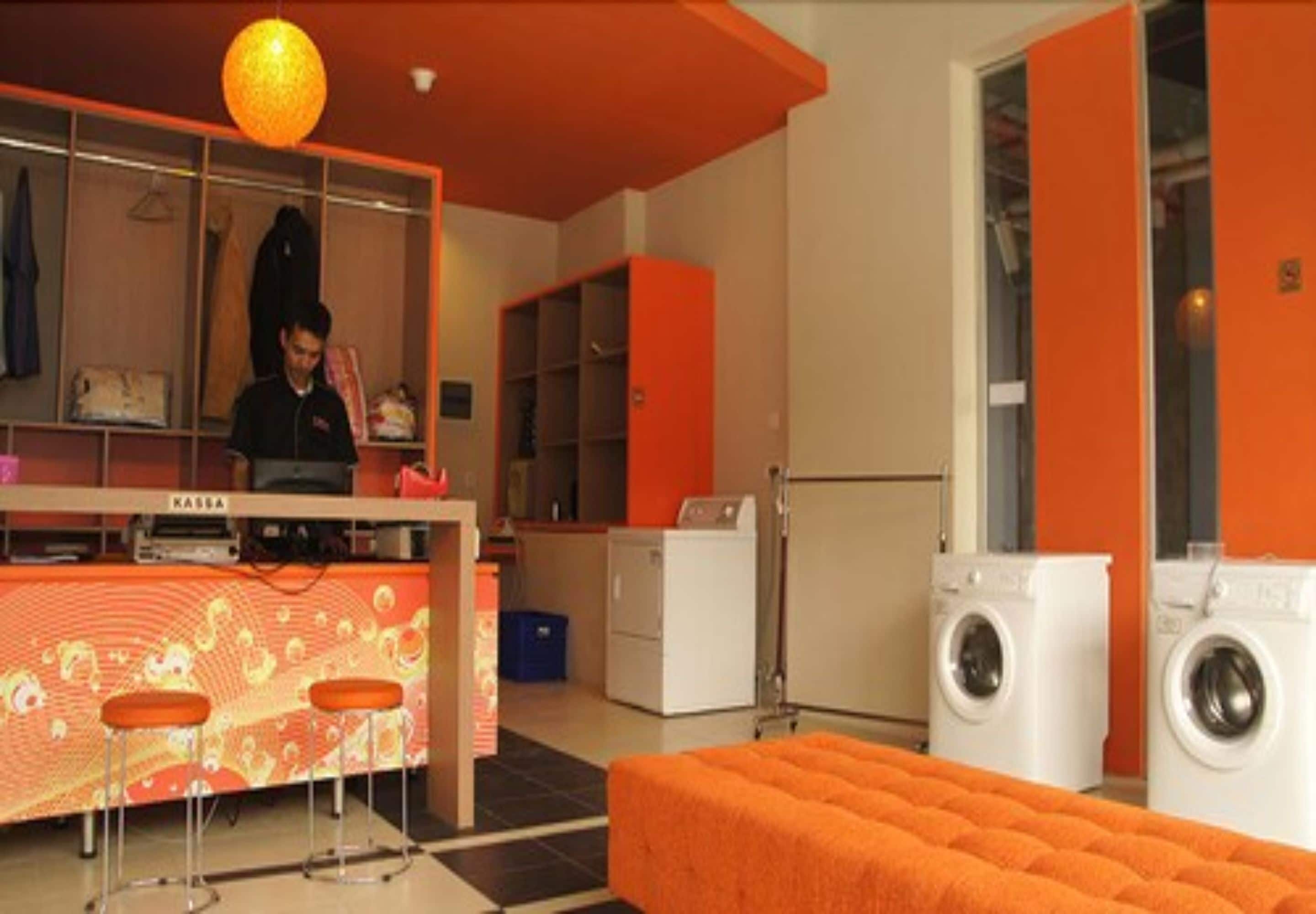 A room with a kitchenette, a bar area with stools, a washing machine, and a large orange ottoman.