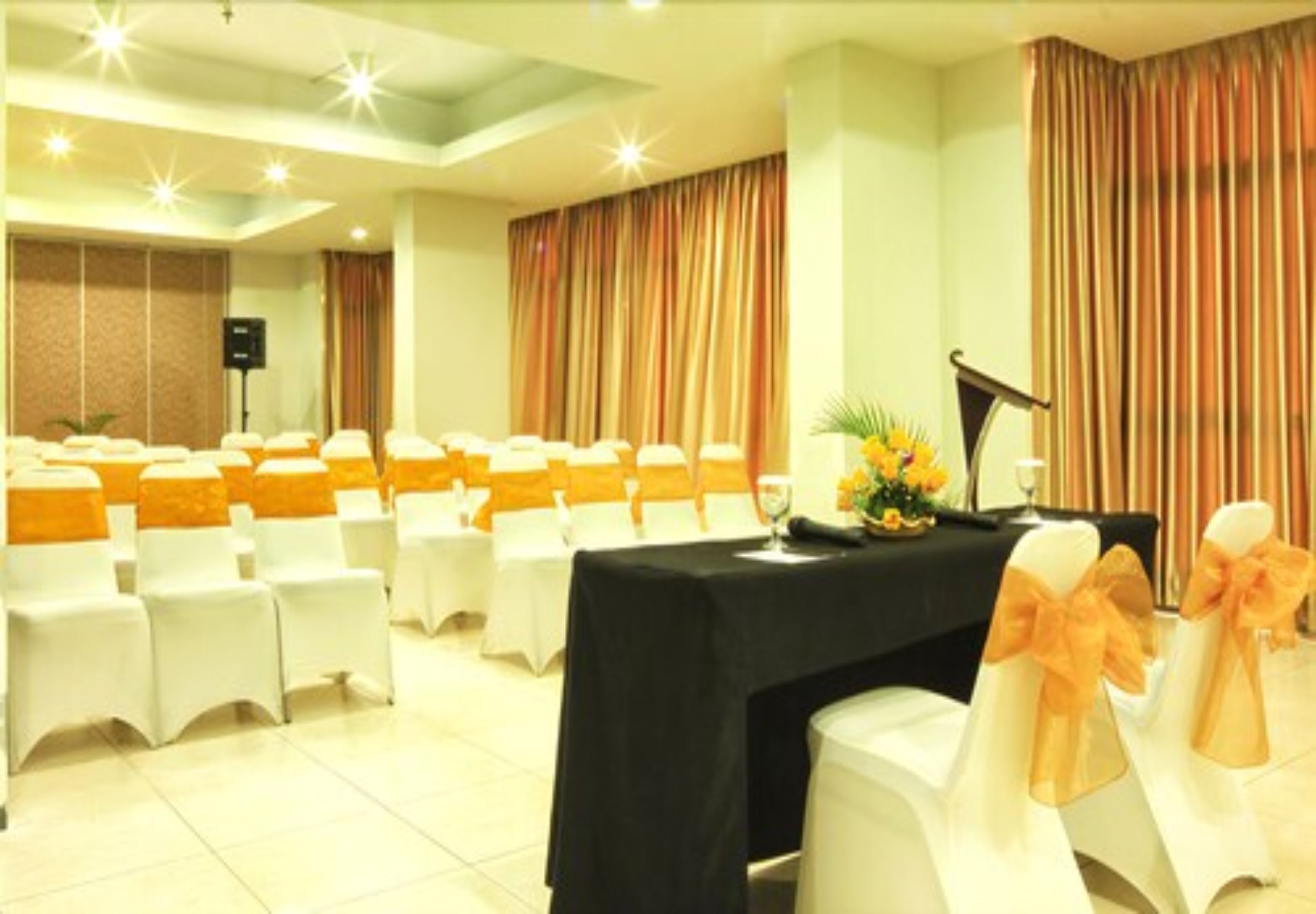 A conference room with rows of white chairs, a black table with a microphone, and curtains.