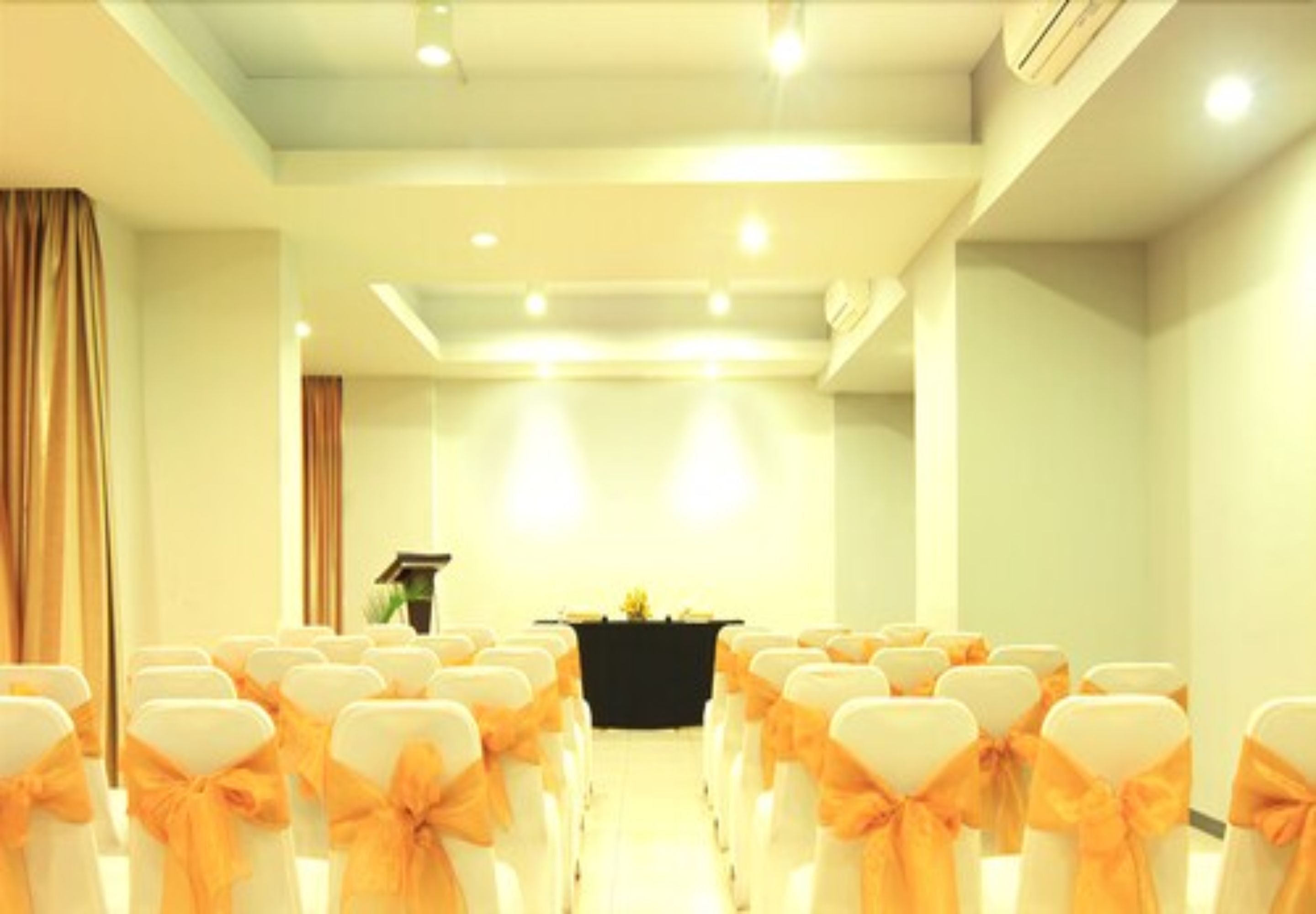 Une salle de conférence avec des rangées de chaises blanches ornées de rubans orange, un pupitre et une table sur laquelle est posé un vase de fleurs.