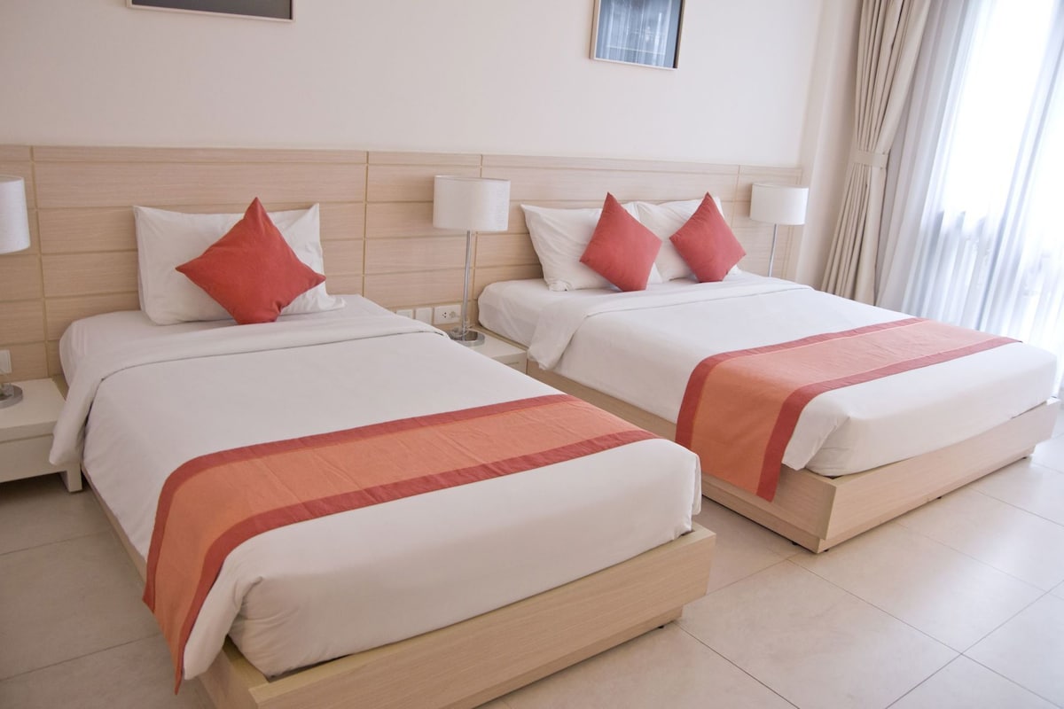 A hotel room with two beds, each with a white mattress and red accent pillows, and a wooden headboard.