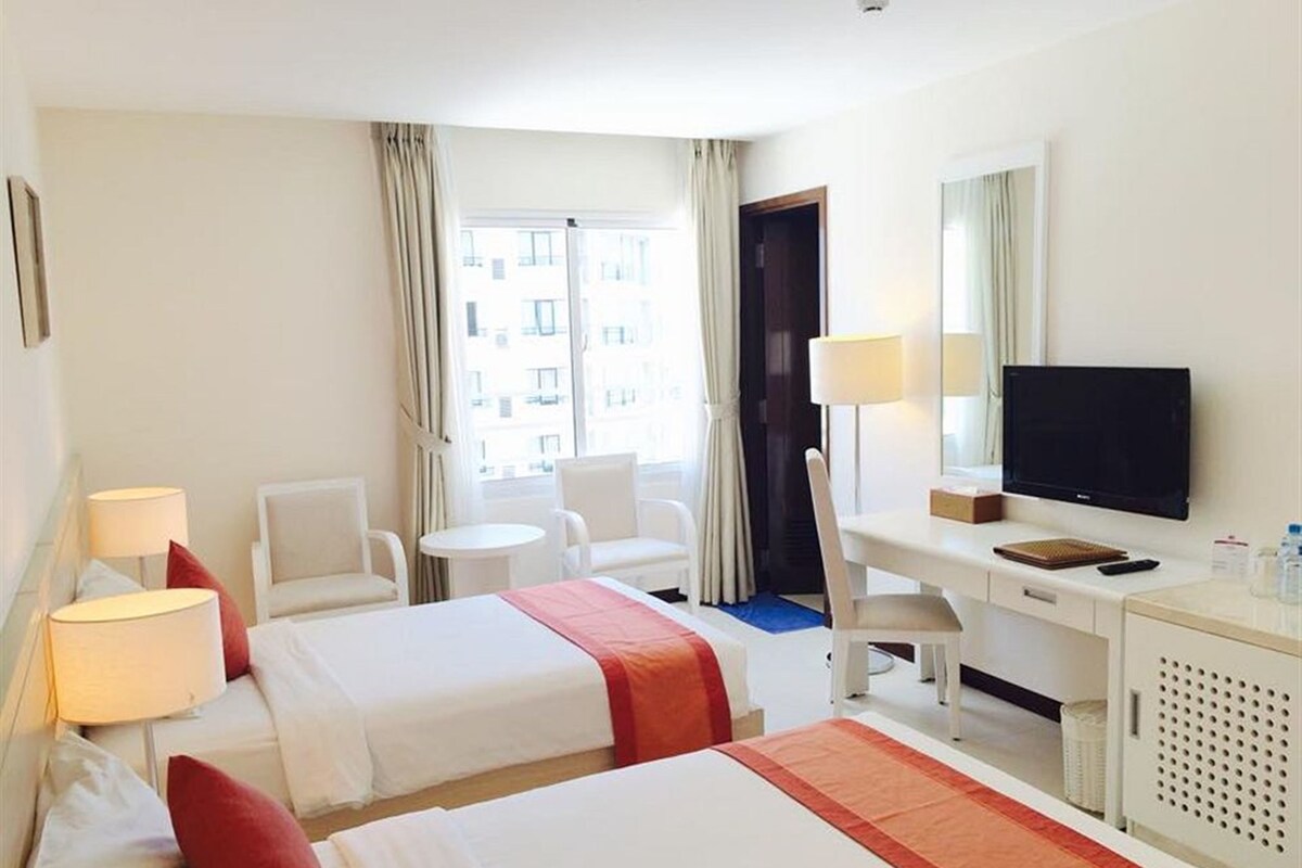 A hotel room with two beds, a desk with a TV, a chair, a small table, and a window with curtains.